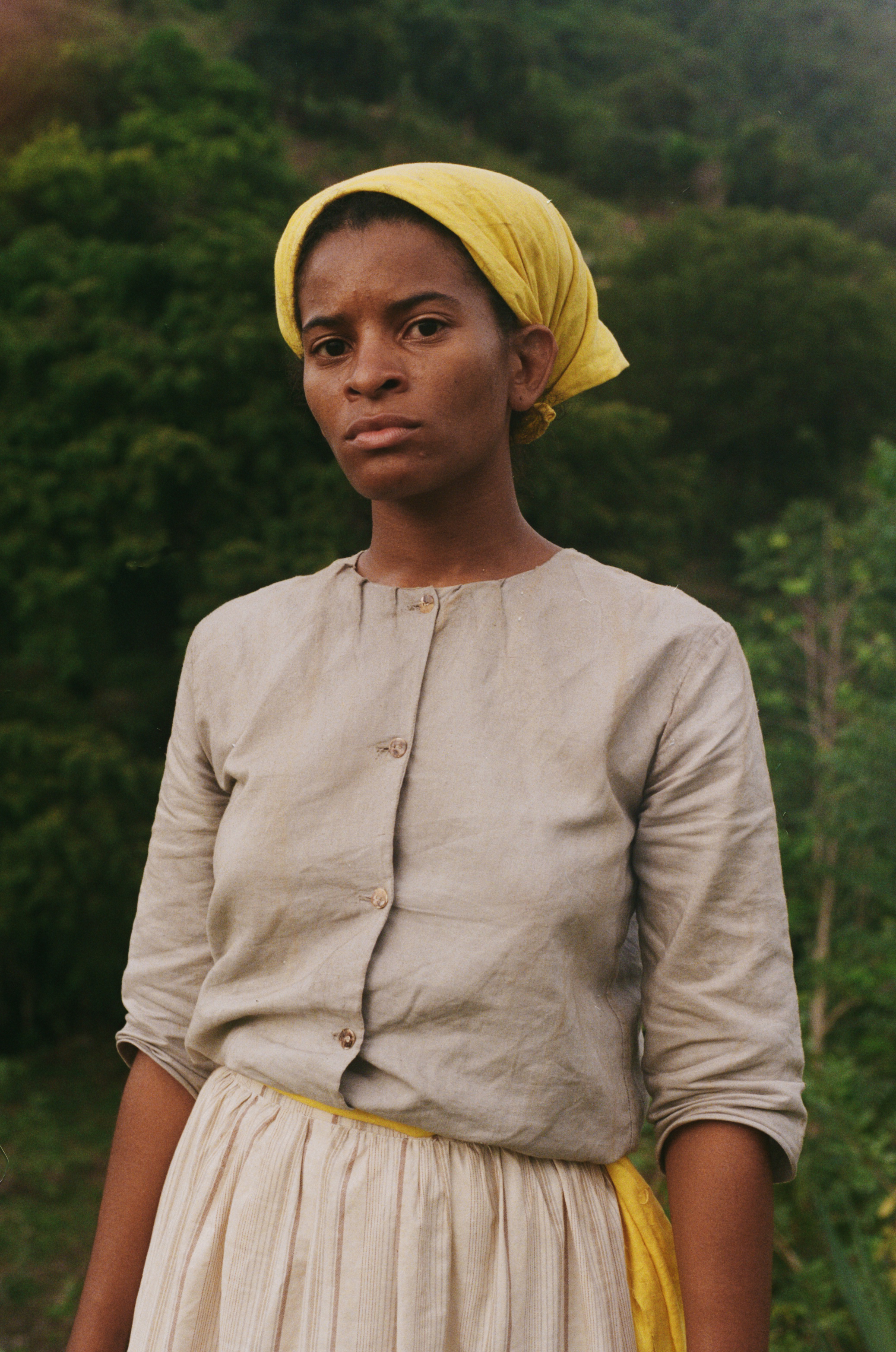 Image Description: A photo of a young woman wearing a course tan dress with a yellow belt and matching yellow head covering. She stands in front of a series of trees and stares into the camera.