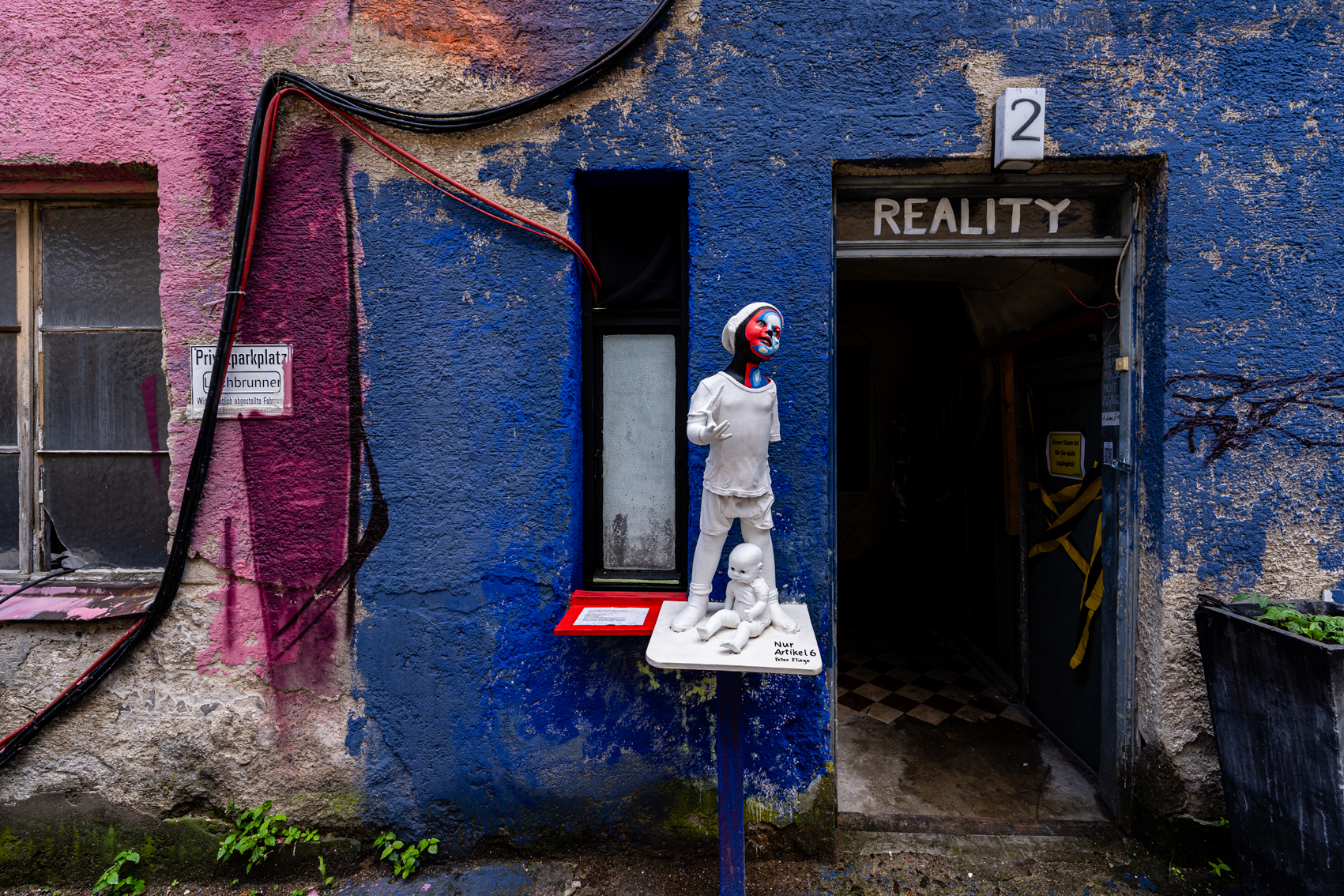 Bild 1 Nur Artikel 6: Links vom Eingang in das Haus Reality 2 steht auf einer wei&szlig;en Holzplatte auf H&uuml;fth&ouml;he eine wei&szlig; bemalte Kinderfigur in Lebensgr&ouml;&szlig;e. Ihr rechter Arm ist angewinkelt, die Finger sind gespreizt und zeigen nach vorne. Der Kopf ist leicht zur Seite geneigt und ihr Blick nach oben gerichtet. Ihr Gesicht ist Rot, blau, wei&szlig; bemalt. Die Figur tr&auml;gt eine wei&szlig;e M&uuml;tze. Zwischen ihren Beinen ist eine weitere Skulptur. Ein Baby, das komplett wei&szlig; bemalt wurde. Es blickt nach links unten.