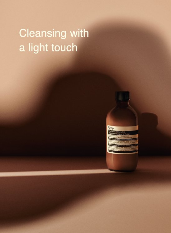 A stylized shot of the Gentle Facial Cleansing Milk bottle, with light illuminating the product label. Copy reads: Cleansing with a light touch.