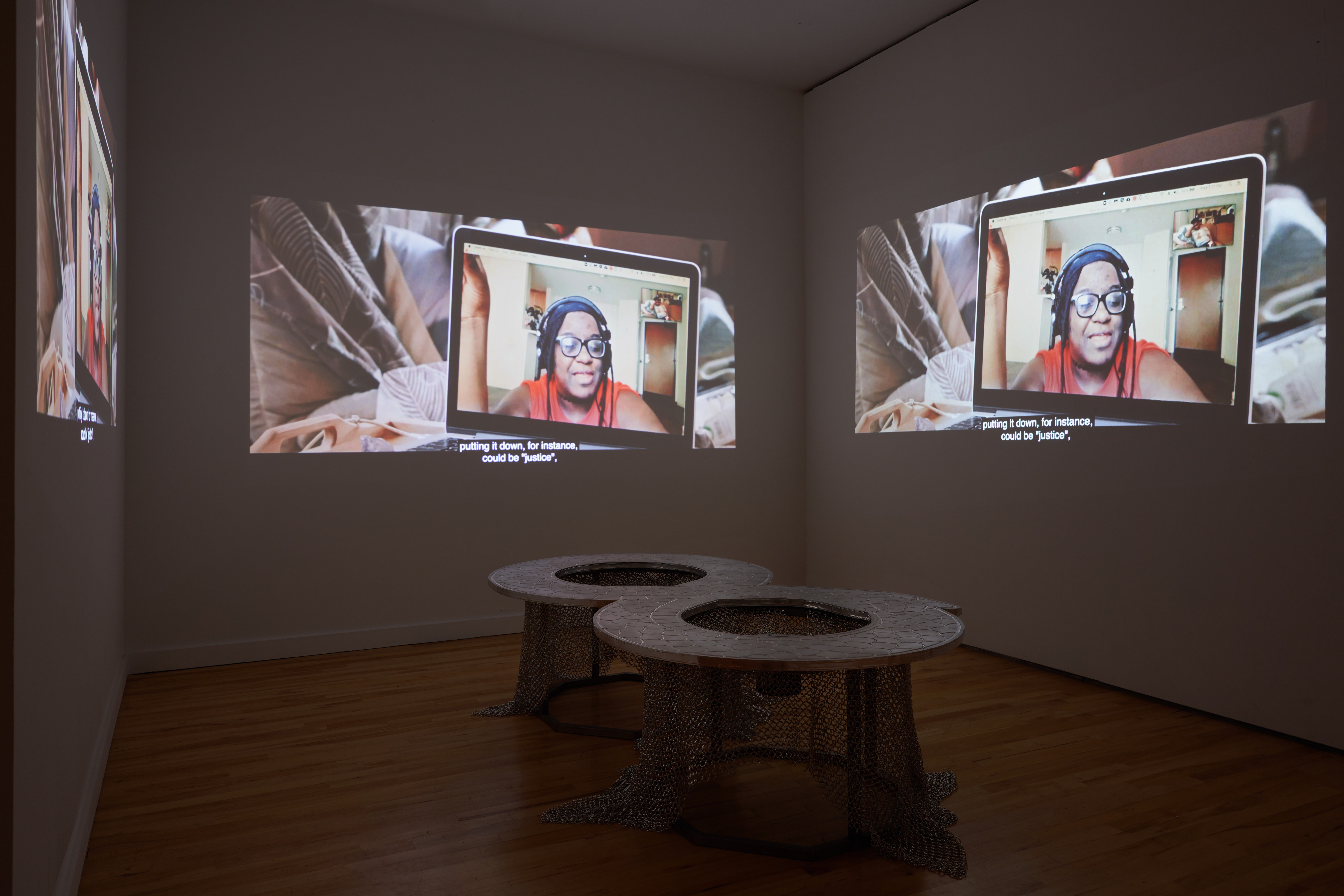 A dimly lit room with three copies of the same projection on three adjacent walls that show a video call on a laptop, with a low table-like sculpture in the center of the room.
