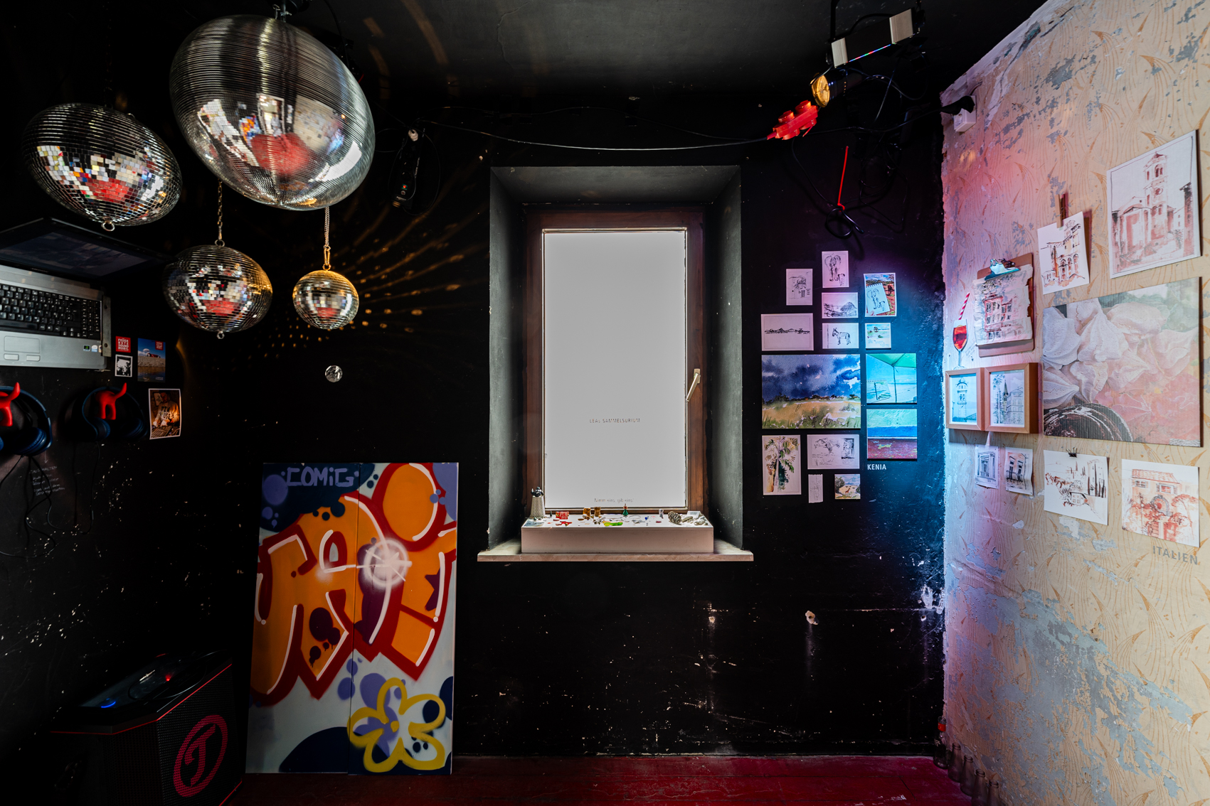 Bild 3 zu IMpressions | IMpressed | IMperfect: Blick vom Eingang des Raumes auf die gegenüberliegende Wand mit einem Fenster mittig der Wand. Links des Fensters steht der zweite Teil des Graffitis. Von der Decke hängen vier verschieden große Diskokugeln. An der linken Wand befindet sich eine Hörstation, sowie ein Laptop.