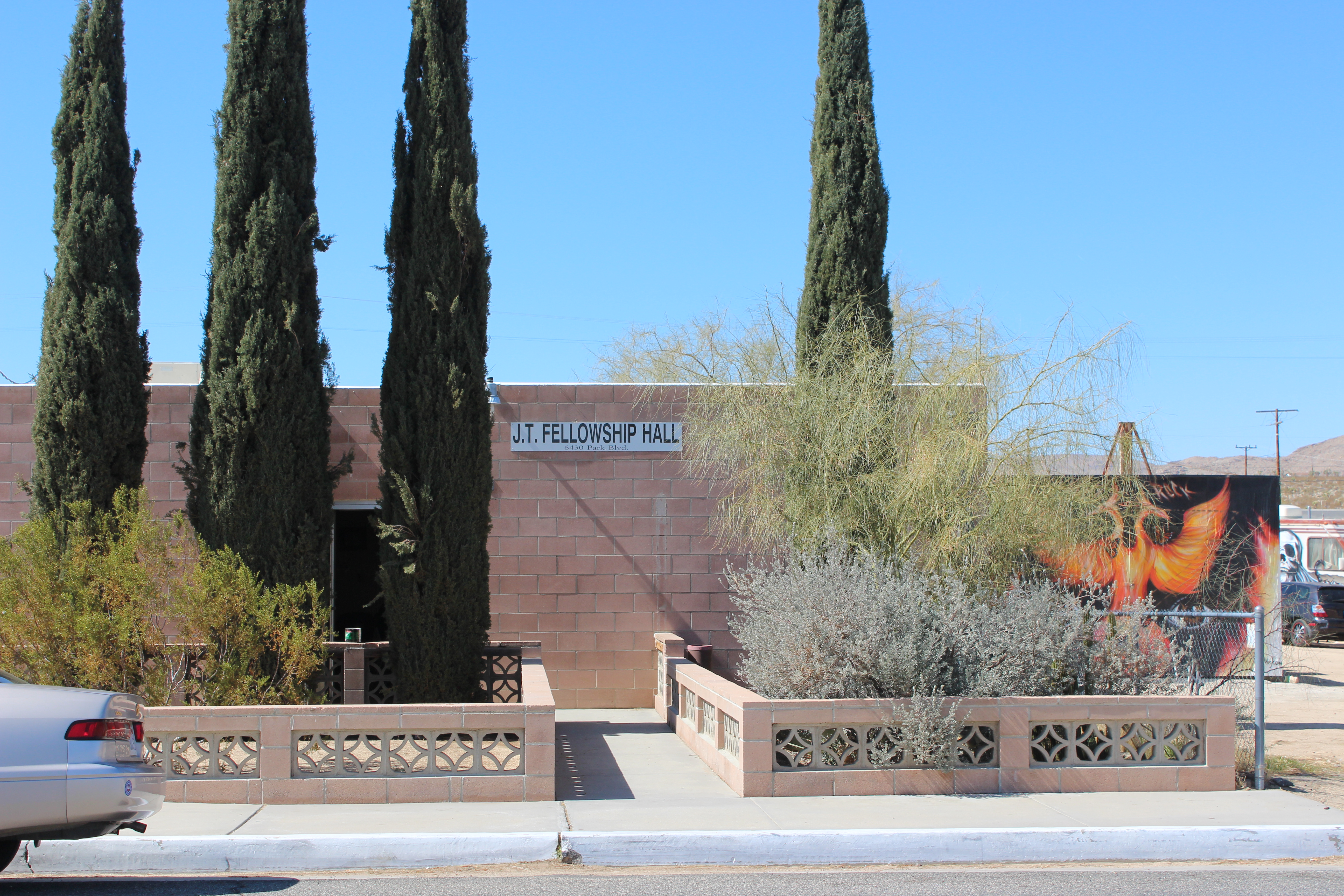 J.T. Fellowship Hall with breezeblock front wall, Palo Verde, Creosote, and Cypress landscaping. A mural of a flaming bird to the side.