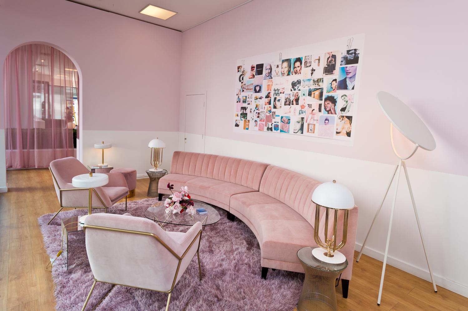 Glossier pop-up interior with curved pink sofa, velvet chairs, mood board wall, and soft pink decor.