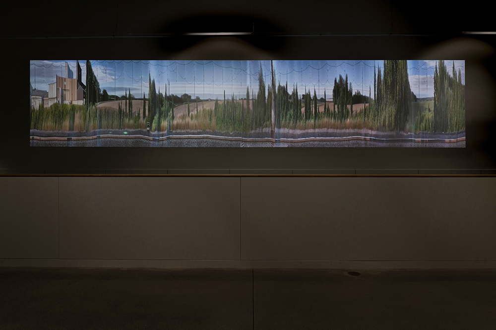 A video projection of an impressionistic landscape of a road, trees, buildings, and the sky in a dark, indoor space.