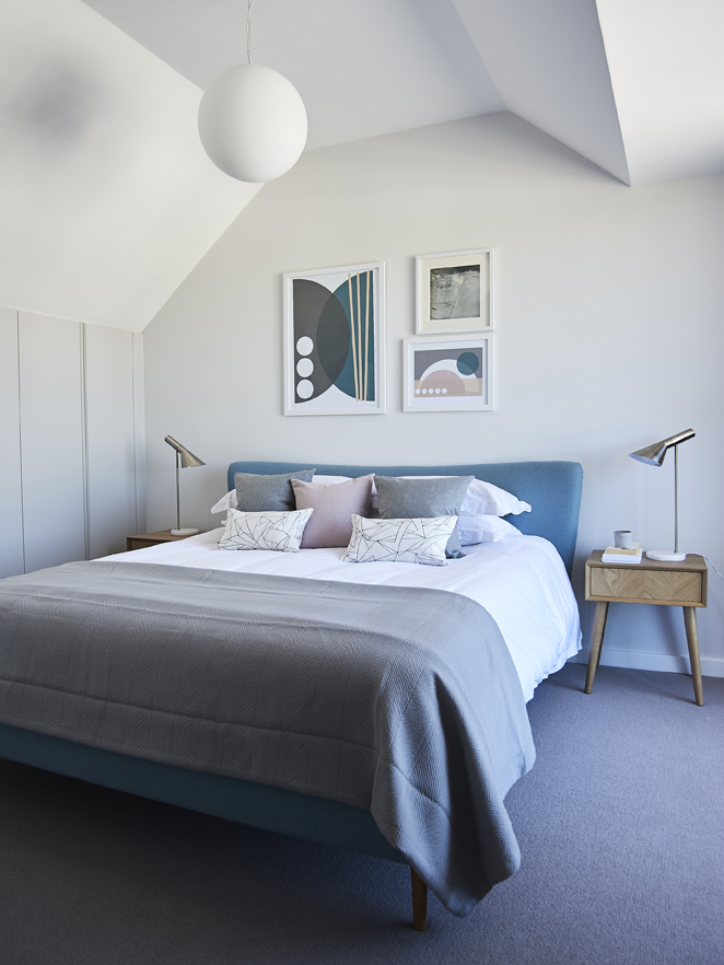 Modern bedroom with blue double bed with grey, pink and white soft furnishings. Abstract art on wall and two side tables.