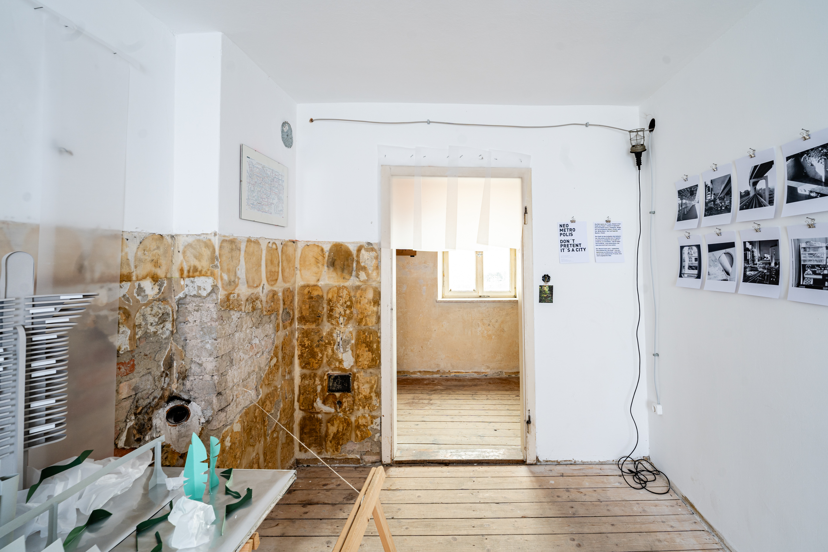 Bild 2  zu Neometropolis: Ausschnitt vom innerem des Raumes, gegen&uuml;ber der T&uuml;r mit Sicht auf den Gang. Zur linken befindet sich das Papiermodell der Stadt und an der rechten Wand auf Augenh&ouml;he die Schwarz-Wei&szlig;-Fotografien. An der linken Wand ist nur die obere H&auml;lfte des Raumes wei&szlig; verputzt, der Rest des Gem&auml;uers ist sichtbar, der Raum hat einen Holzdielenboden.  