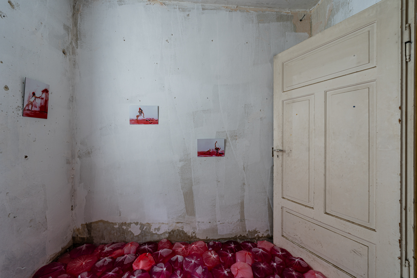 Bild 1 zu RED: Ein wei&szlig;er Raum mit ge&ouml;ffneter wei&szlig;er Holzt&uuml;r rechts. Am Boden befinden sich in verschiedenen Rott&ouml;nen mit Farbe gef&uuml;llte durchsichtige Beutel. An den W&auml;nden h&auml;ngen vereinzelt Fotografien, die die K&uuml;nstlerin bei vergangenen Kunstperformances zeigen. 