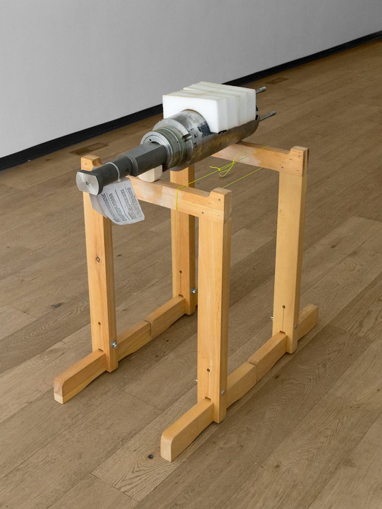 A photo of the heavy steel part known as a plunger/barrel assembly that came from the Atlantic Conveyor. It is sitting on a wooden stand in the gallery. 