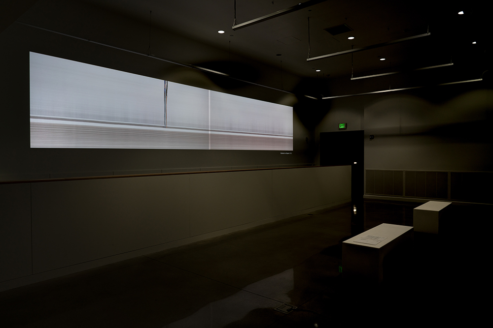 A video projection of two elongated figures and two benches placed in front of the projection in a dark, indoor space.