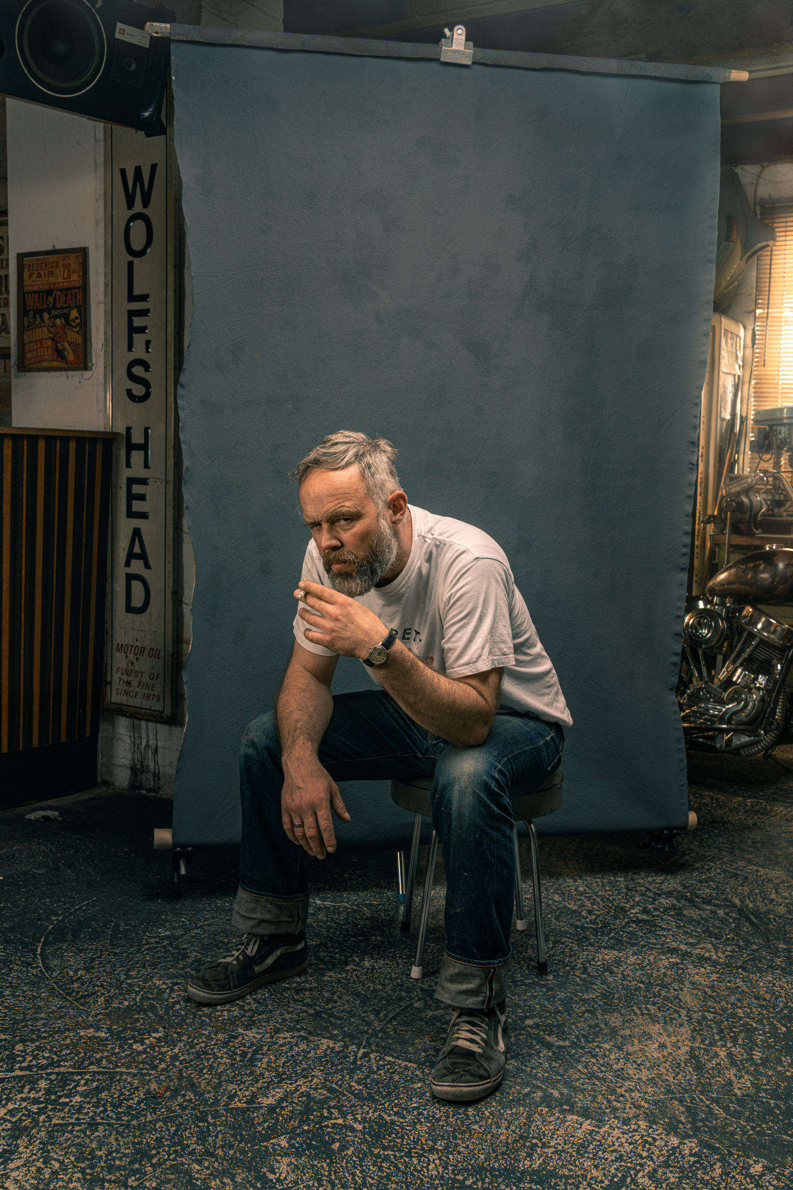 Environmental Portrait aus der Serie The Workshop. Alte Werkstatt, warme Atmosphaere. Ein Mann mit Zigarette sitzt auf einem Hocker vor einem Stoffhintergrund.