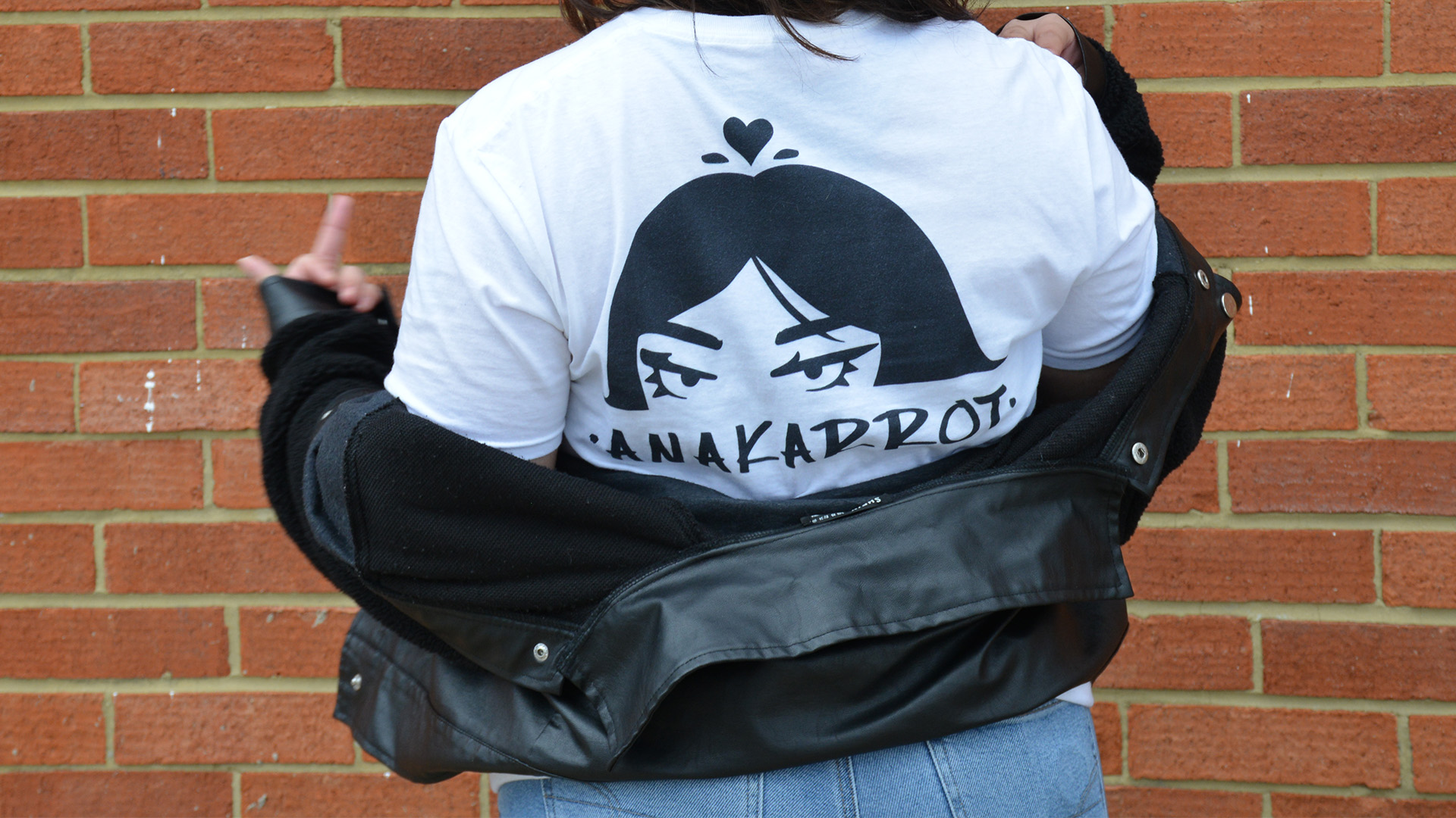 The Back of a girl who is wearing a shirt that was designed for the brand anakarrot. The design is the logo of the brand which is a drawing of a girl with a flower in the shape of a heart on top of her head. And the letters ANAKARROT written right under.