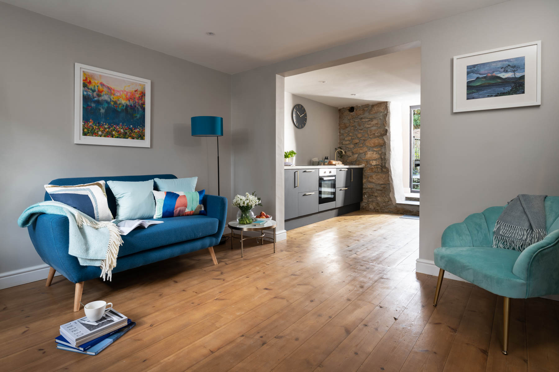 Lounge with teal sofa, arm chair and floor lamp. Side table on timber floor. Kitchen visible through opening.