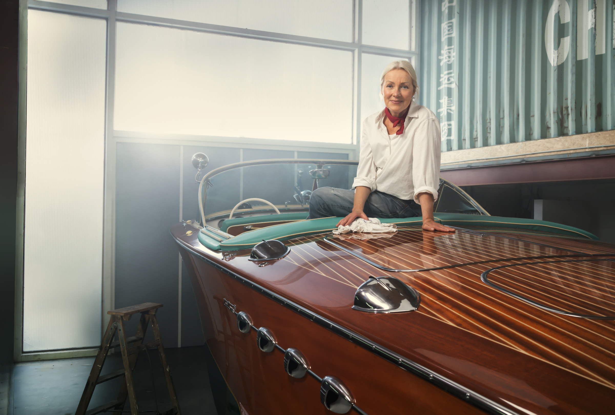 Environmental Portrait aus der Serie The Workshop mit einem alten Riva-Boot. In einer Bootswerkstatt sieht man eine aeltere blonde Frau in Jeans und weißer Bluse sitzend im Riva Holzboot.