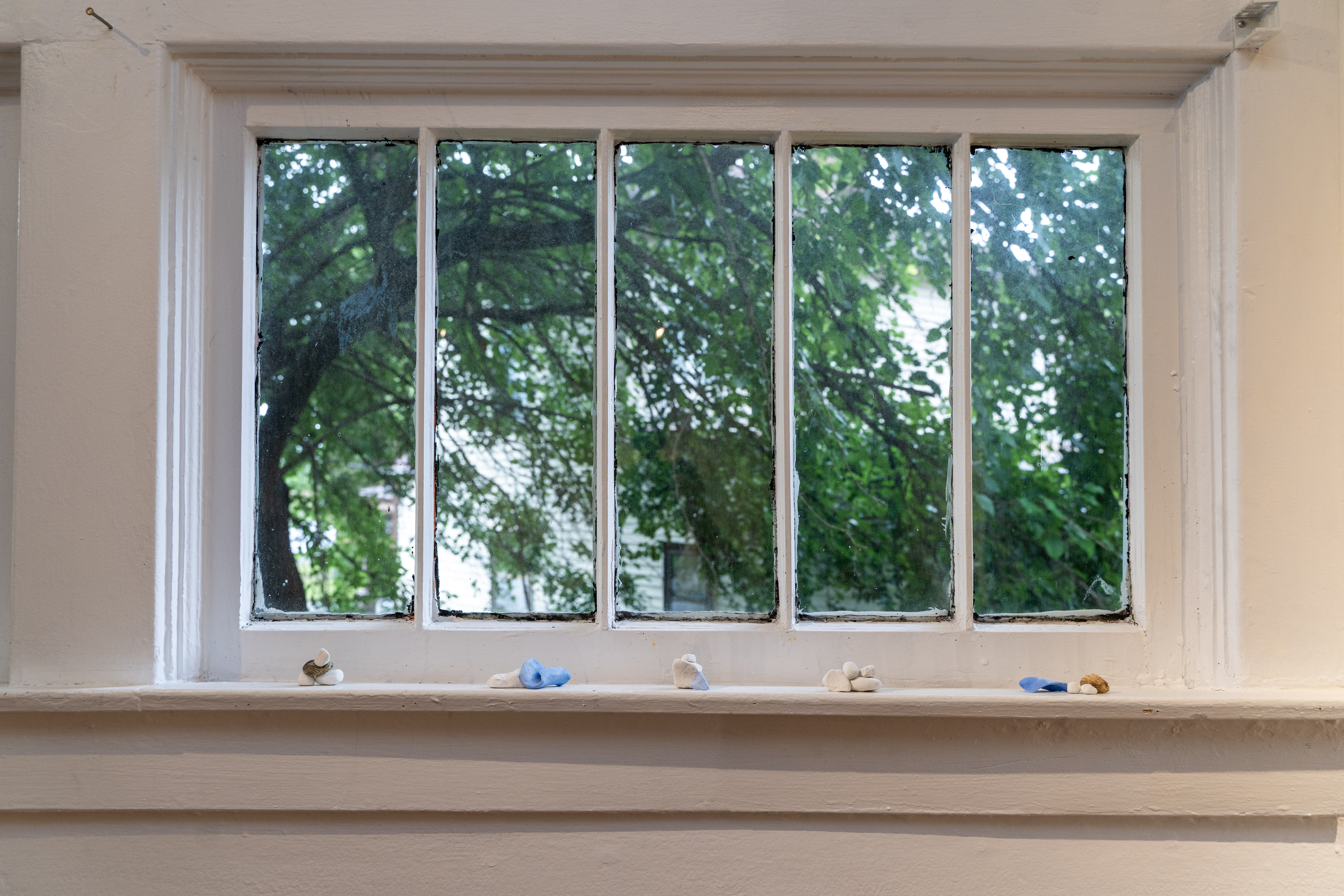 An interior view of a window with five vertical panes of glass, its frame and sashes painted white, overlooking a tree and a white house outside. On the ledge of the window sit five small clusters of objects that look like rocks or lumps of clay, mostly white with some blue and brown pieces.