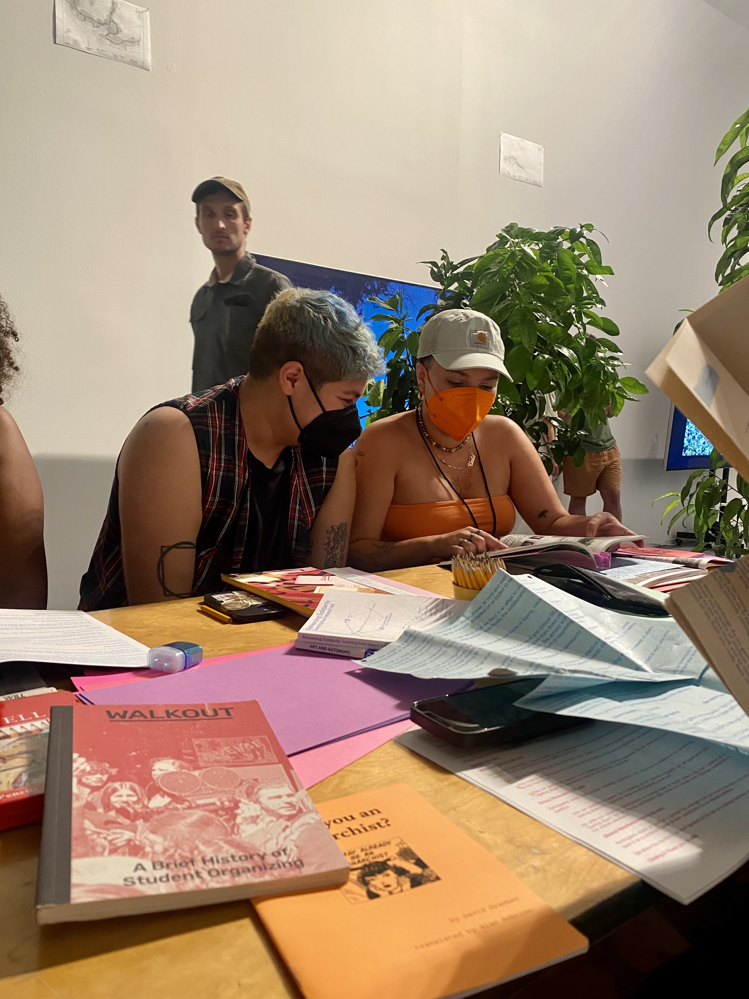 Two participants lean into looking at a book together on top of a table strewn with printed material. One of the participants wears a bright orange mask matching their tube top.