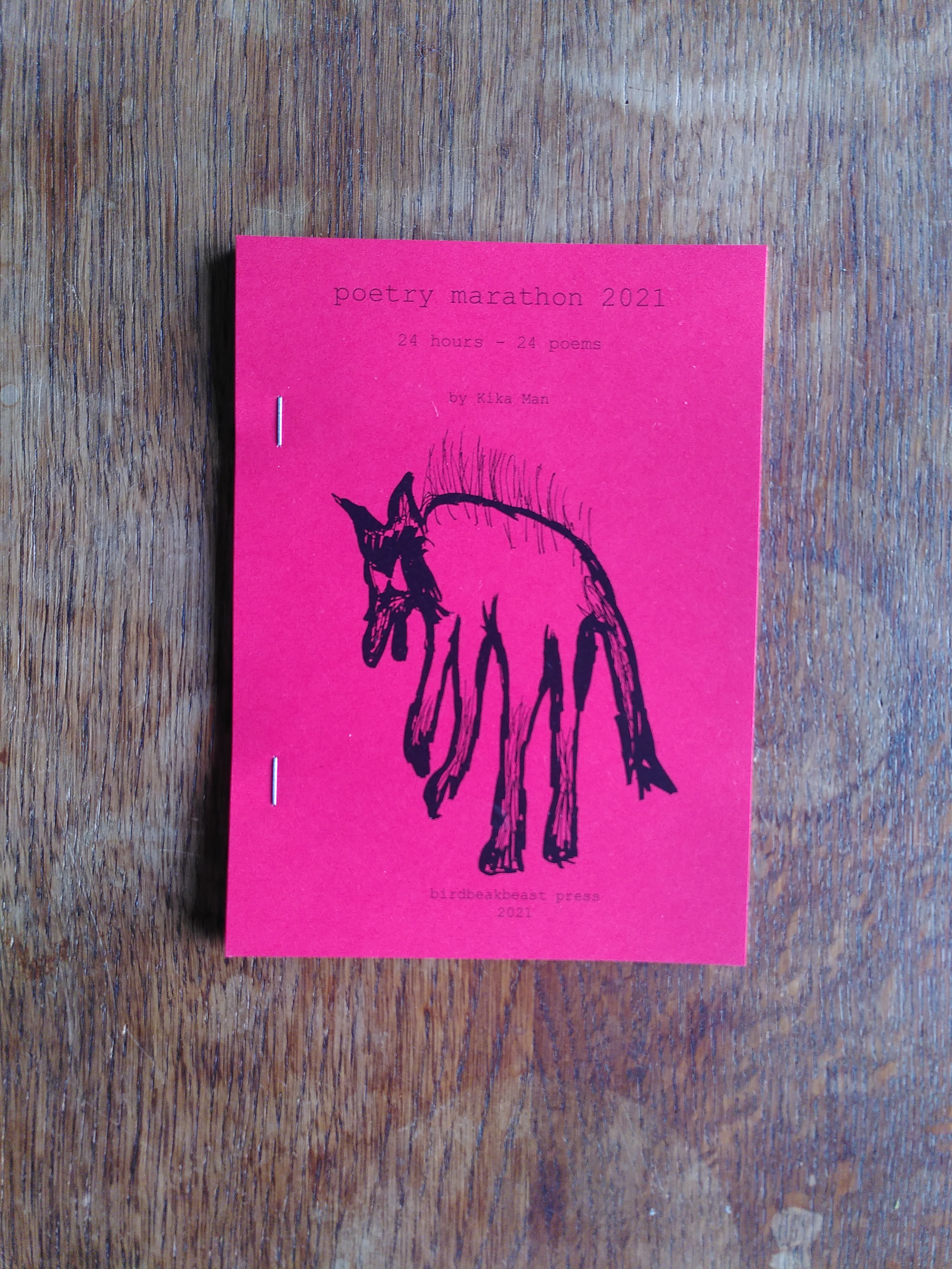 cover of 'poetry marathon 2021' by Kika Man, a dark-red zine, on a wooden table. a black dog is shown, above which it reads 'poetry marathon 2021, 24 hours - 24 poems, by Kika Man'.
