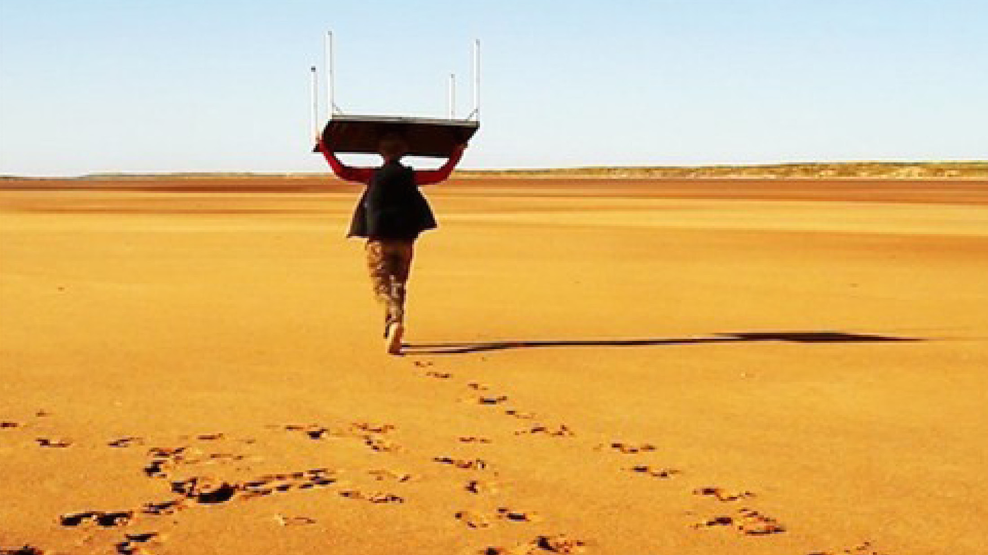 ID: View of a flat desert plane. A person in the mid-ground carries a card table over their head. They are walking away from the camera. The foreground shows a line of footprints behind the person. In the foreground is a mass of footprints circling around an area.