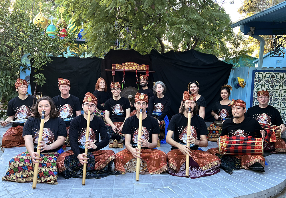 A group of 14 musicians sits on a stage outdoors for the camera. They are either next to their instruments or holding them.