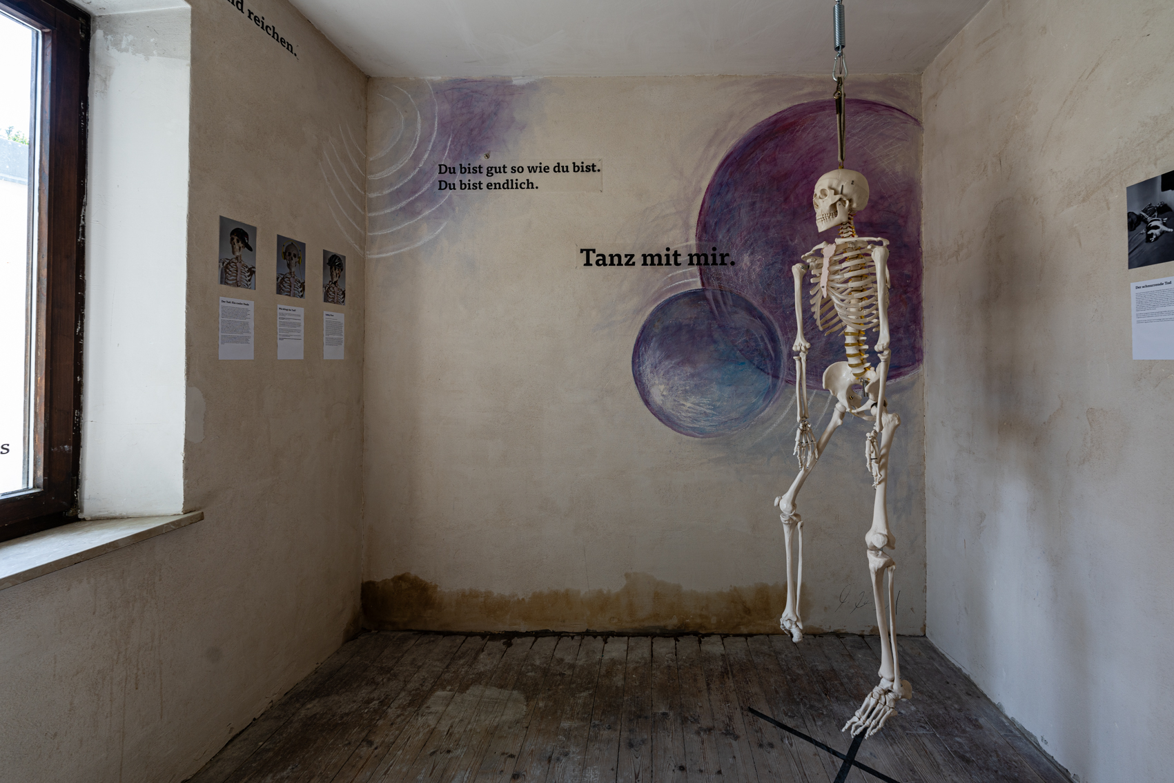 Bild 1 zu Augsburger Totentanz: Ein quadratischer Raum mit einem Fenster zur linken. An der Decke h&auml;ngt ein menschengro&szlig;es k&uuml;nstliches Skelett. An der Wand gegen&uuml;ber rechts im Eck sind zwei verschieden gro&szlig;e violette Kreise gemalt. Auf der Wand steht mit Druckbuchstaben "tanz mit mir" und "du bist gut so wie du bist, du bist endlich".
