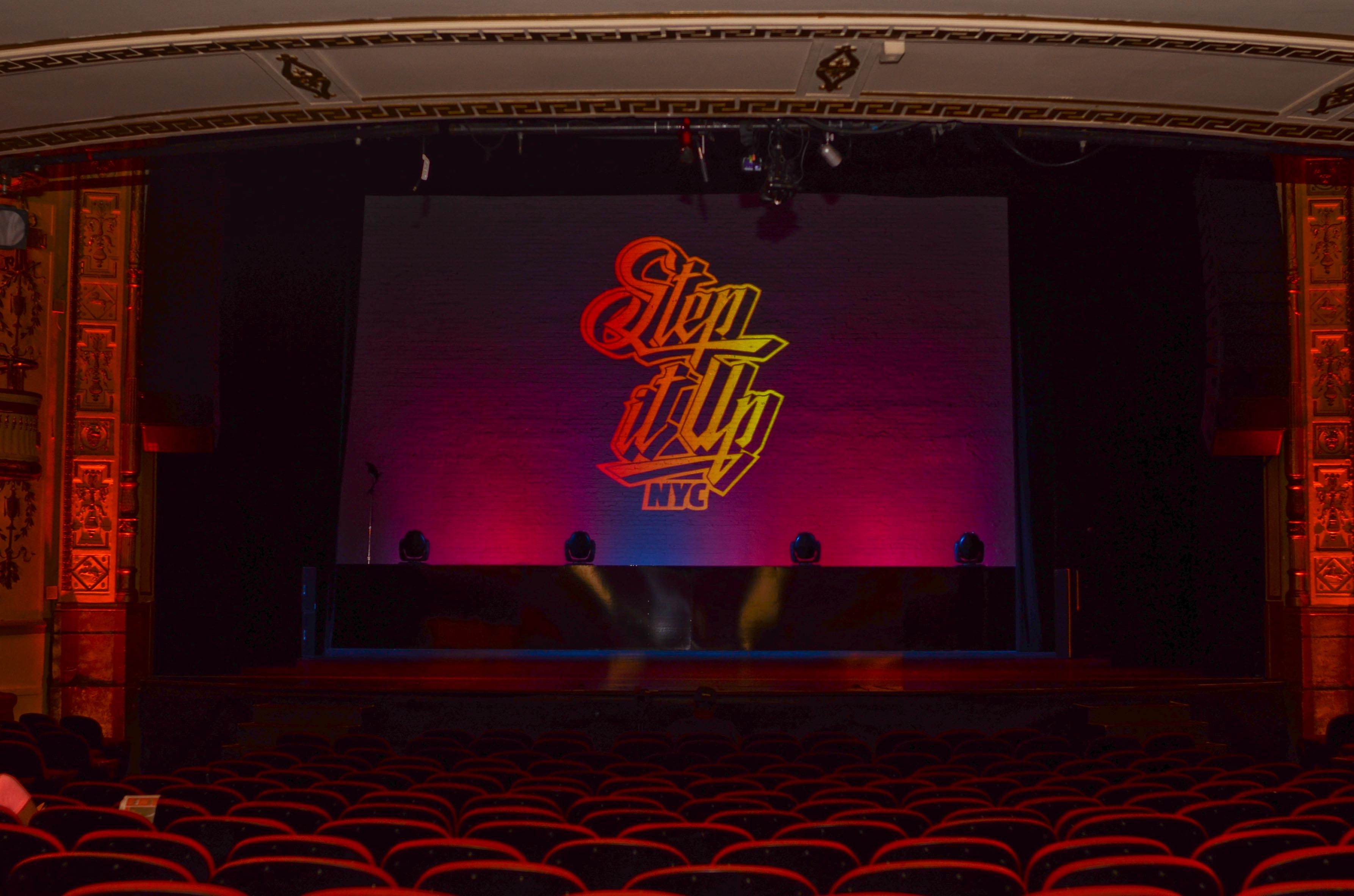 The gradient full color Step It Up NYC logo projected on stage inside an empty Harlem Apollo Theater.