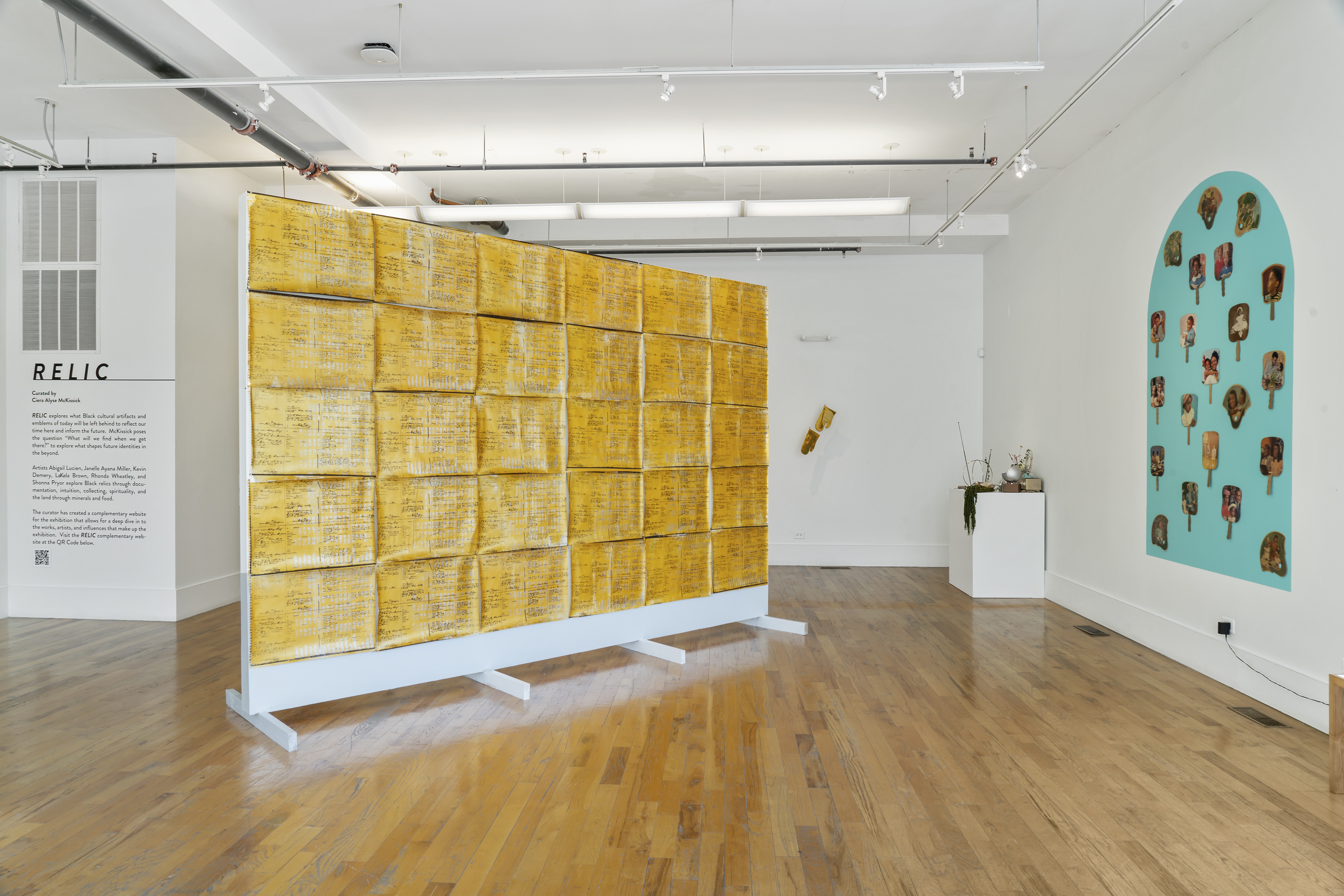 Installation of an art exhibition in a white wall gallery space with wood floors. On the left is a very large floor standing sculpture with a grid of shiny yellow papers with very small text all over. On the right is a display of over 20 church fans hung in front of a blue painted shape on the wall.