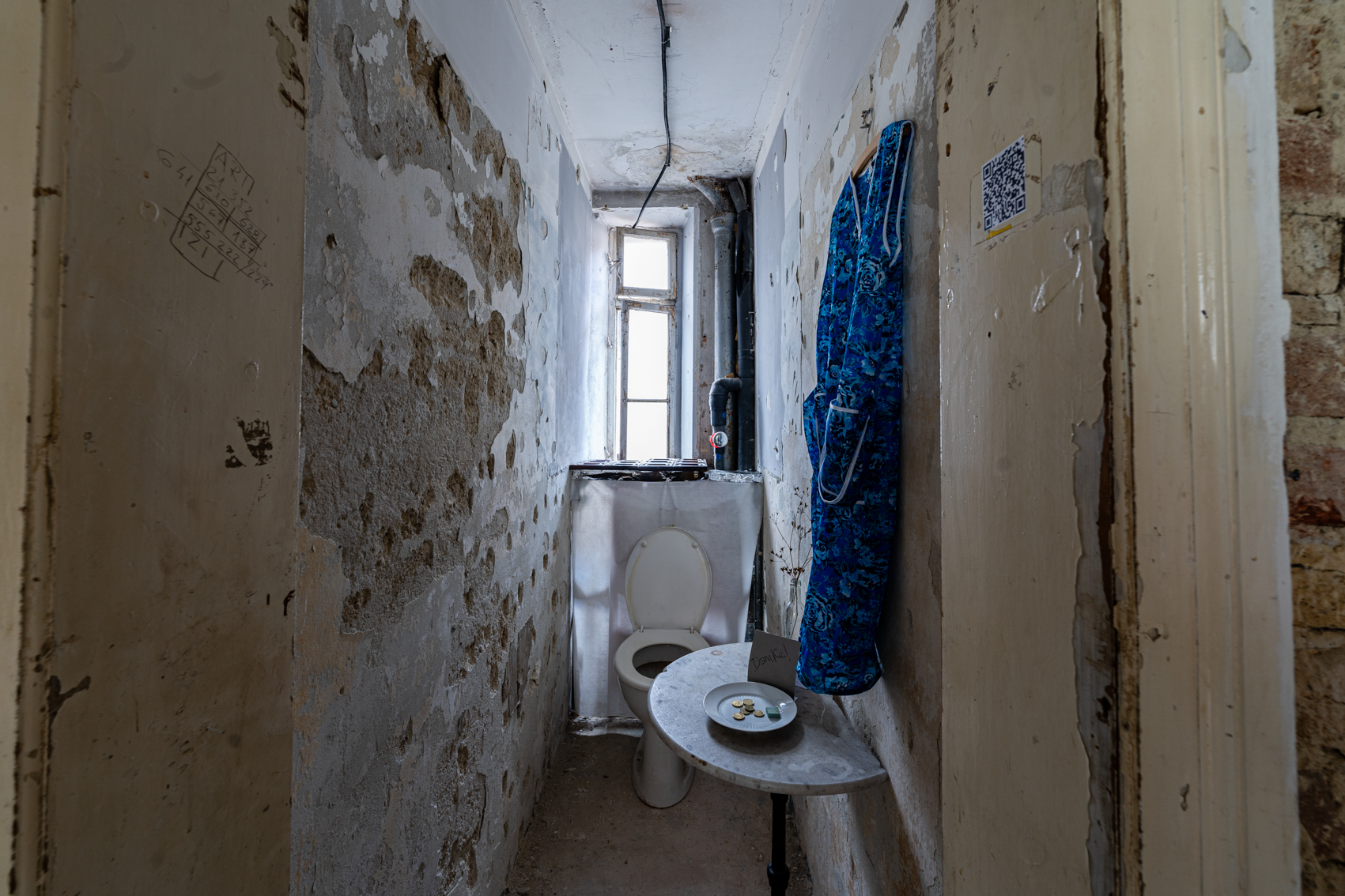 Bild 1 zu Klolekte: Ein schmaler Raum mit einem Klo, dahinter ein schmales, leicht ge&ouml;ffnetes Fenster. Die W&auml;nde sind unverputzt, teilweise br&ouml;ckelt das Gem&auml;uer. Rechts an der Wand nahe der Eingangst&uuml;r steht ein halbkreisf&ouml;rmiger Marmortisch mit der geraden Seite zur Wand. Darauf steht ein Spendenteller. Dahinter steht auf einem Schild "Danke!". Rechts davor an der Wand h&auml;ngt ein blau gebl&uuml;mtes Kleid, wie es h&auml;ufig Reinigungskr&auml;fte tragen.