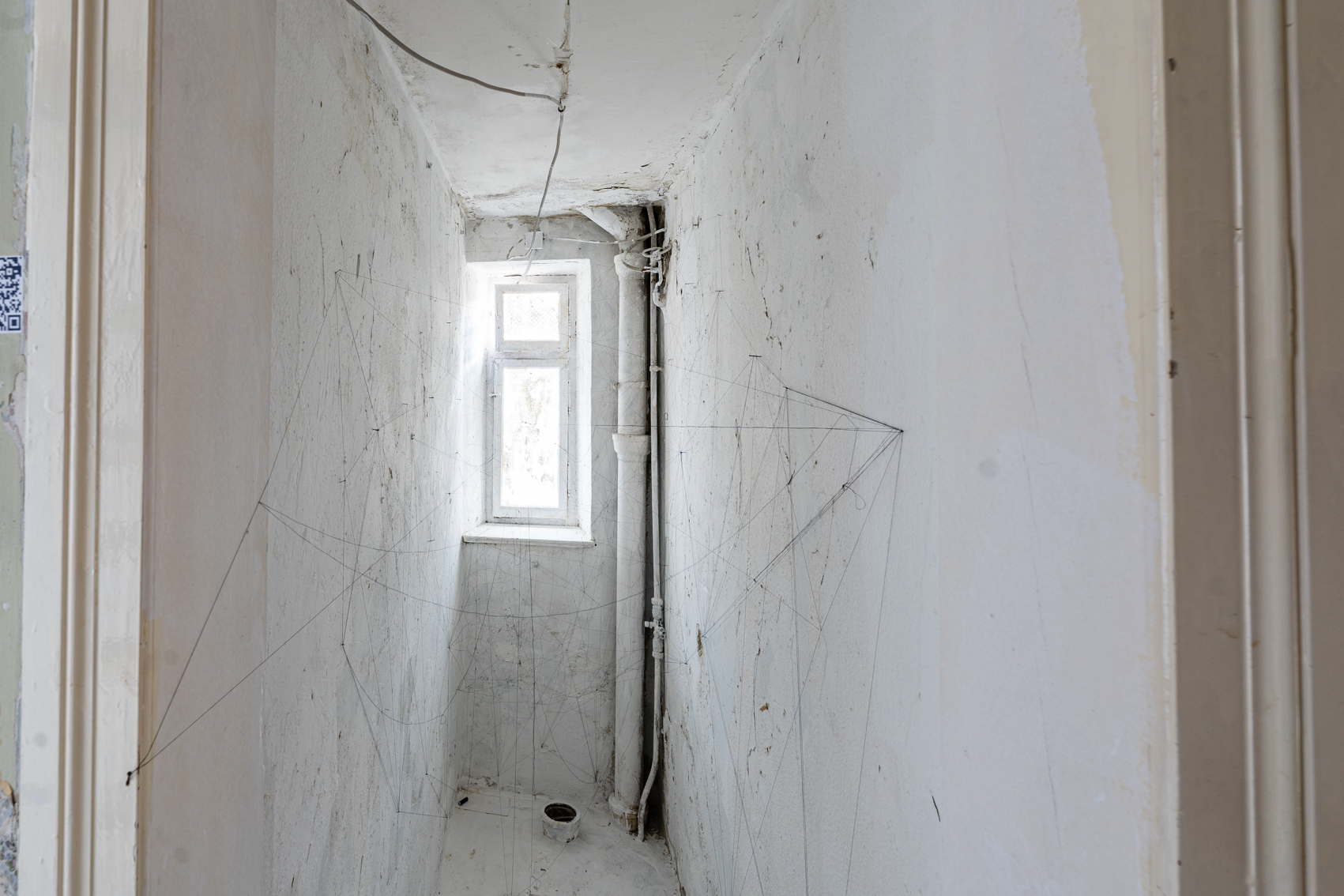 Bild 1 zu Please hold the line: Schmaler wei&szlig;er Raum mit einem kleinen Fenster am Ende. In der hinteren rechten Ecke ein wei&szlig;es Rohr. Zwischen den W&auml;nden sind d&uuml;nne schwarze F&auml;den gespannt, die Spinnennetzen &auml;hneln.