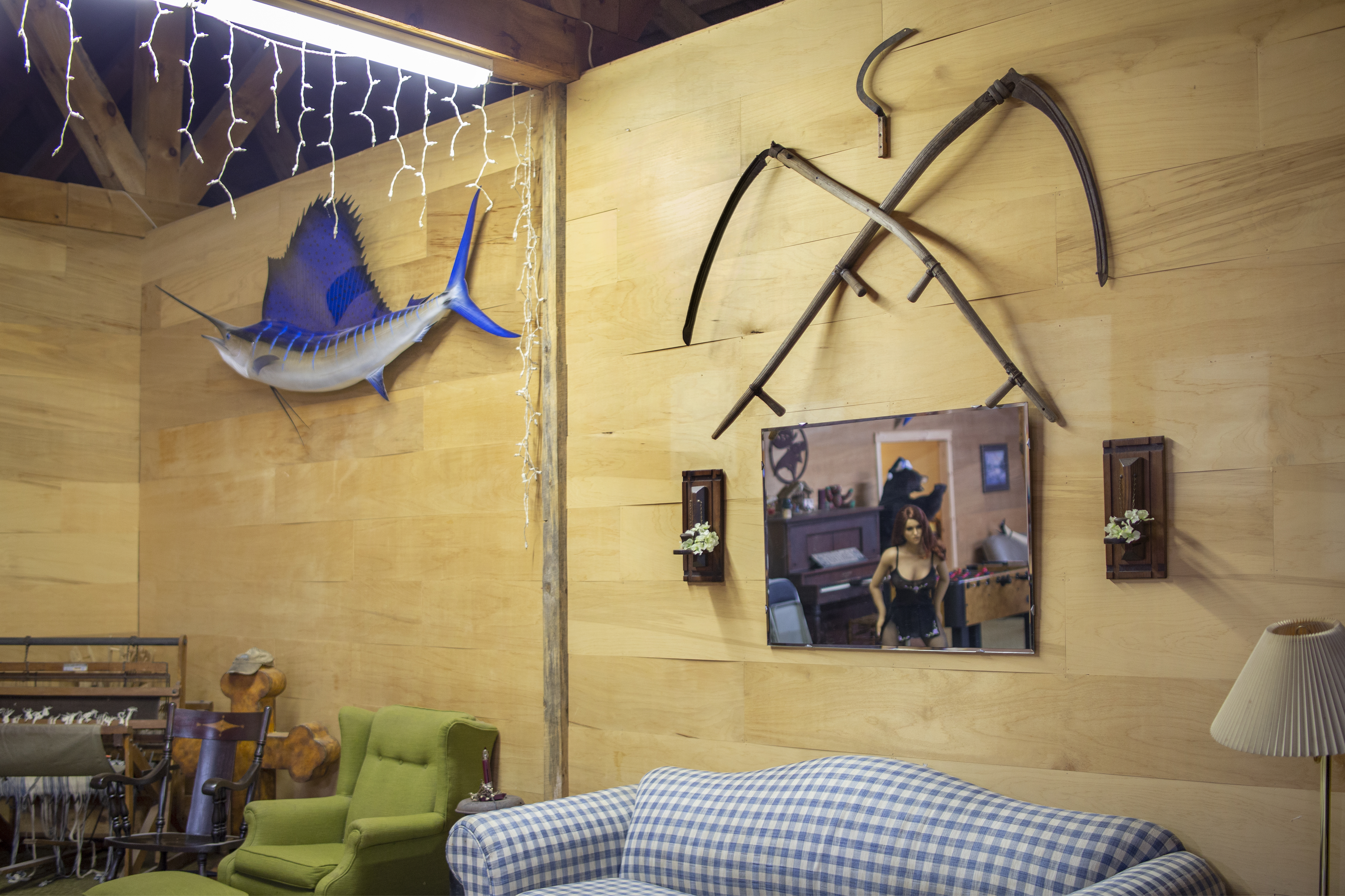 [A corner of a room is lit with a fluorescent light bar and hanging string lights. From right to left, upholstered and wooden seats are aligned against wooden walls. A replica of a blue swordfish hangs above a loom  and next to three scythes: two are arranged in an X shape while a third and smaller scythe hangs above them. Beneath the blades hangs a mirror. In the reflection is white silicone Doll in black lingerie. In her background are similar wooden walls, a piano, a foosball table, and a taxidermied black bear wearing a Santa hat standing on its hind legs in front of a doorway.]