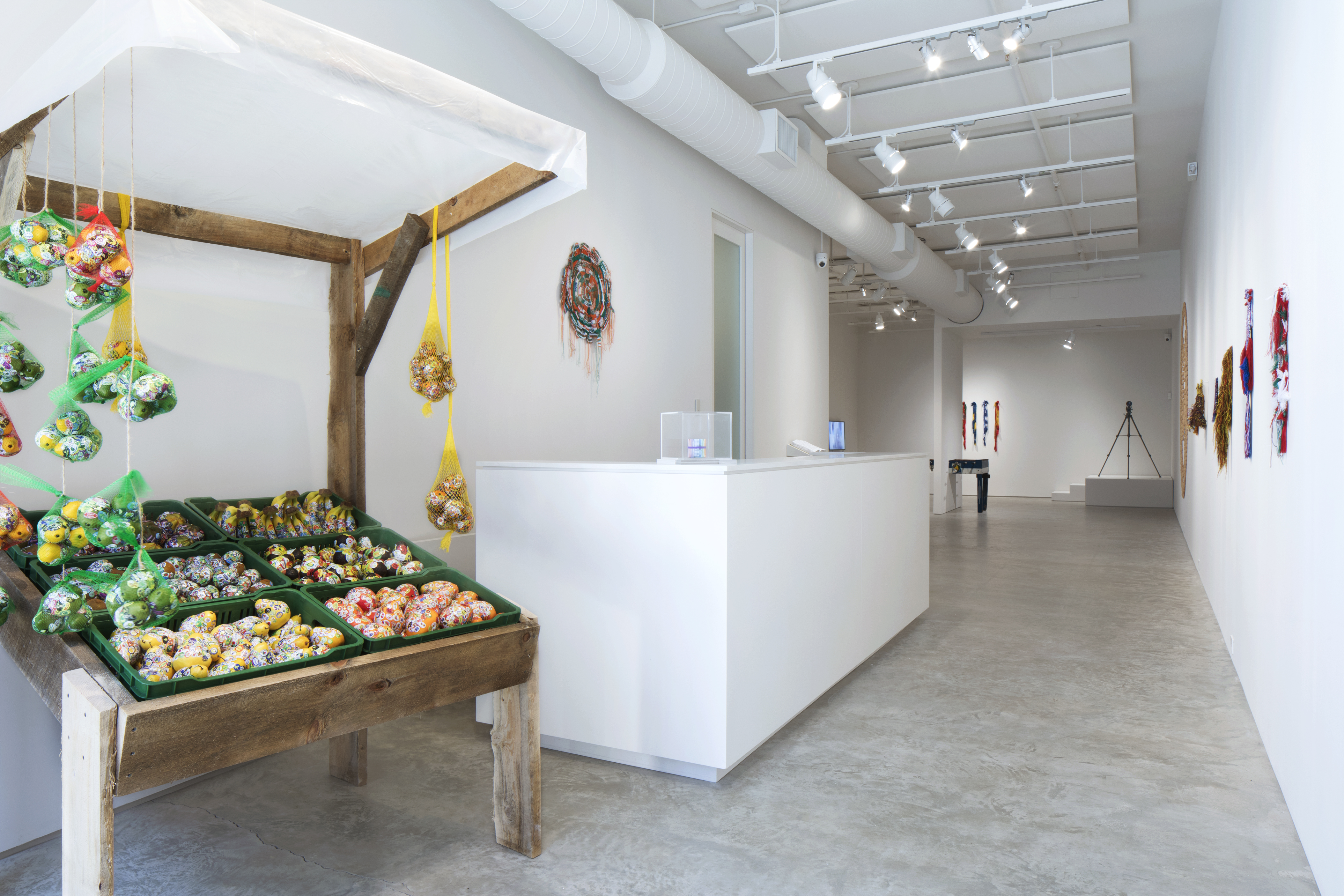 Installation View, Holes in Maps curated by Juliana Steiner. Photographer: Marie Noële Guex. From left to right: Adriana Martinez, Tutti Frutti Market, 2018, plastic fruits, stickers, six plastic crates, wood, mesh, 7' x 4' x 4'. Adriana Martinez, Al Final del Arco Iris, 2015, currencies of Costa Rica, Nicaragua, Venezuela, Chile and Brazil, rubber band, 3" x 3." Sanne Vaassen, FLAGS series, 2018, Rewoven flags.  