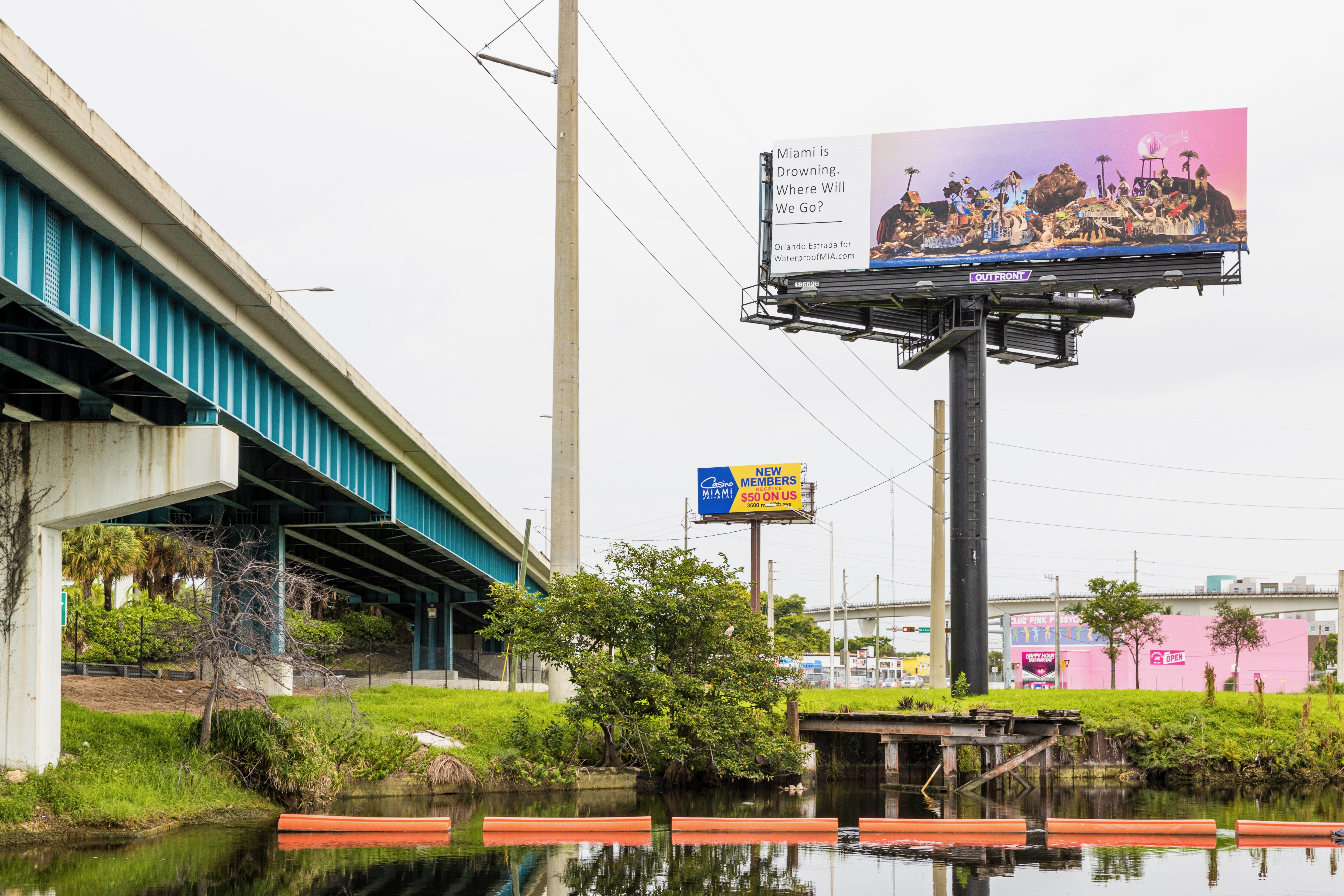 A billboard, resting on the right side of a highway by a canal, depicts an island sunset with a tropical heaven on the left side and a dystopian hell on the right beside a text box that reads in black, “Miami is Drowning. Where Will We Go? Orlando Estrada for WaterproofMIA.com”.
