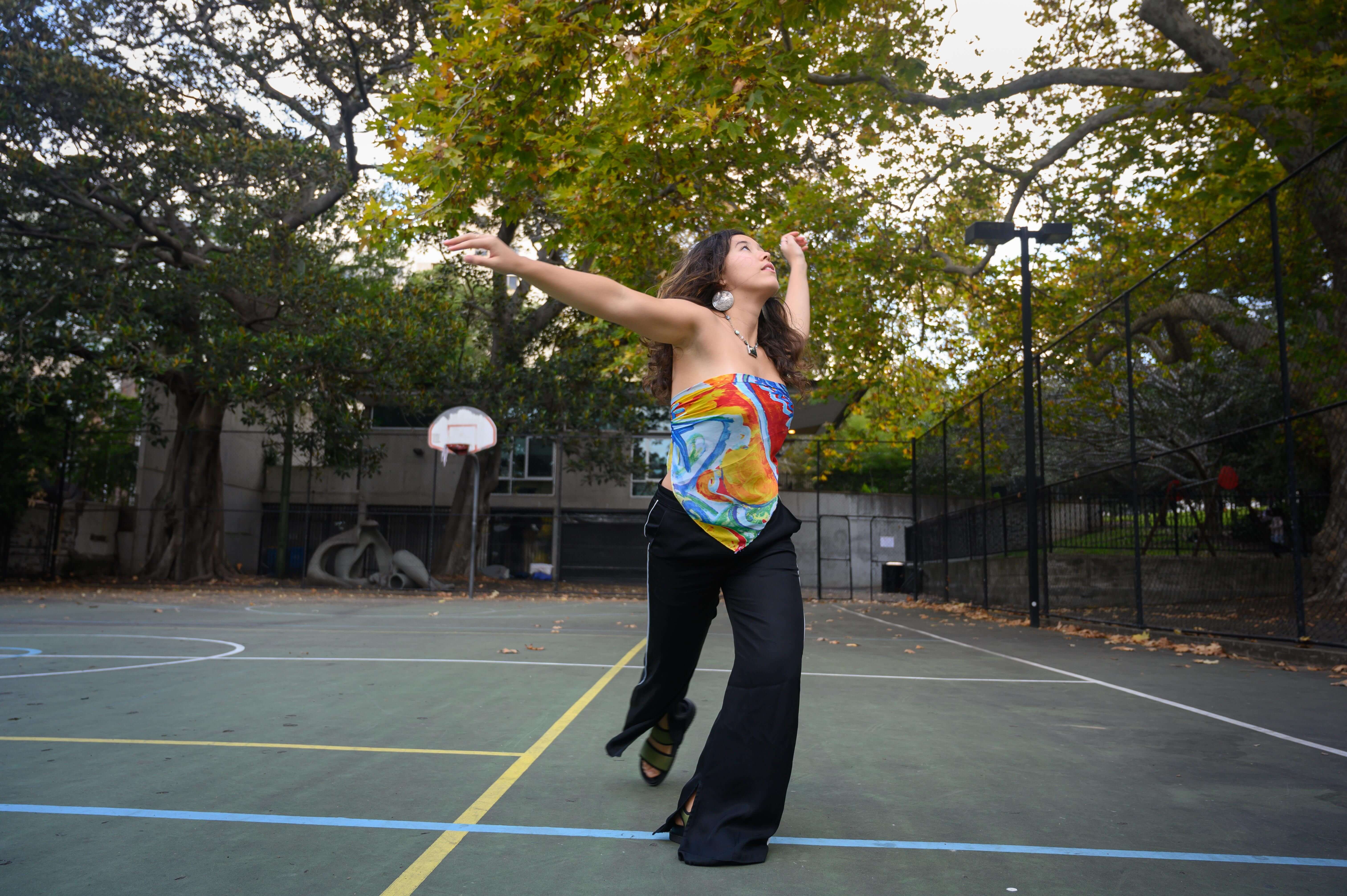 Reina is standing at a basketball court and is poised mid-dance. Her body is expressive and open, her gaze is directed to the sky.