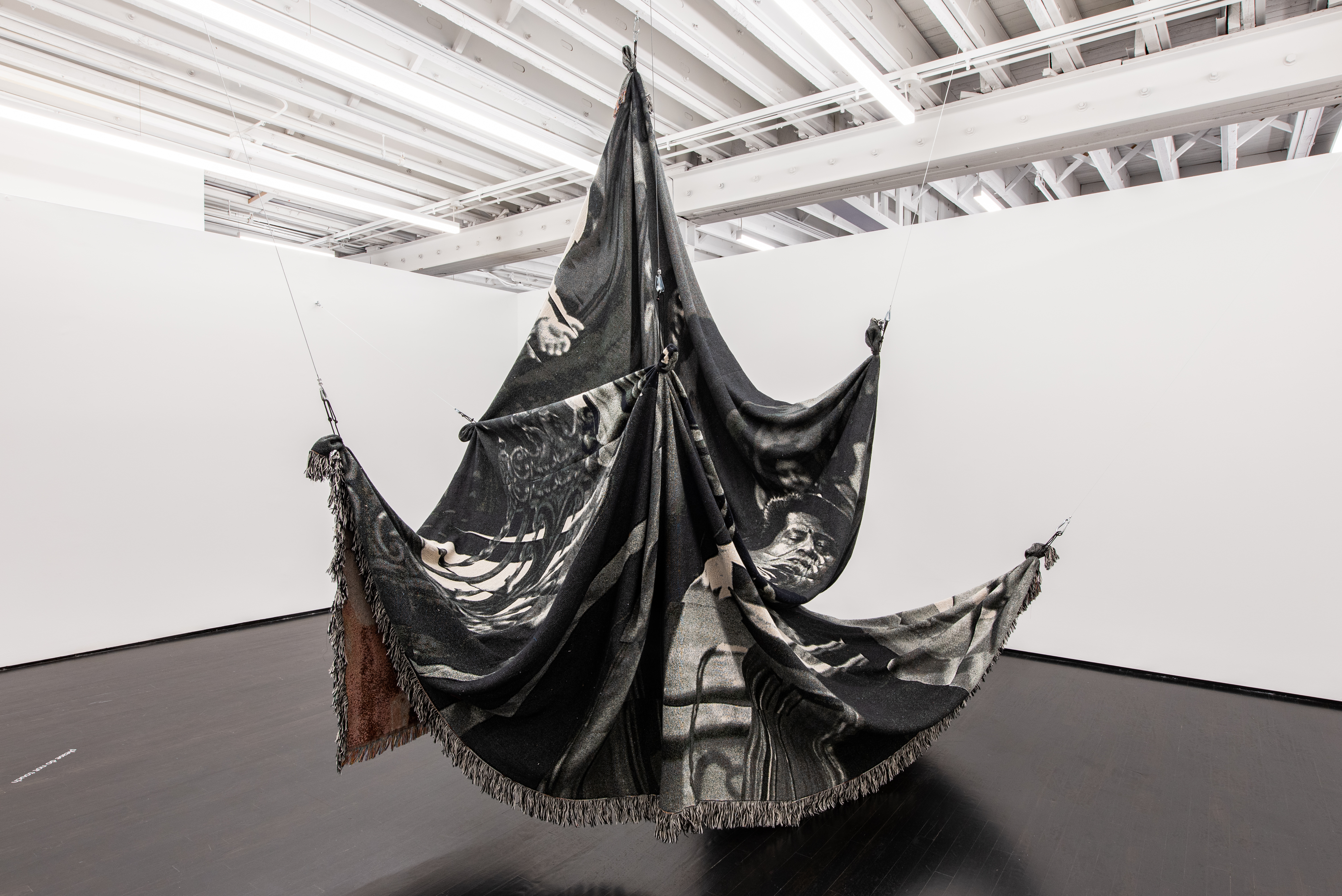 A woven jacquard tapestry hangs suspended in the middle of a white-walled gallery space, draping from wires that stretch to the walls and ceiling at various angles. The tapestry is black and white with an image of James Brown in his role from the 1980 movie The Blues Brothers discernible on the front, and some color visible where the back of the tapestry shows through underneath folds.
