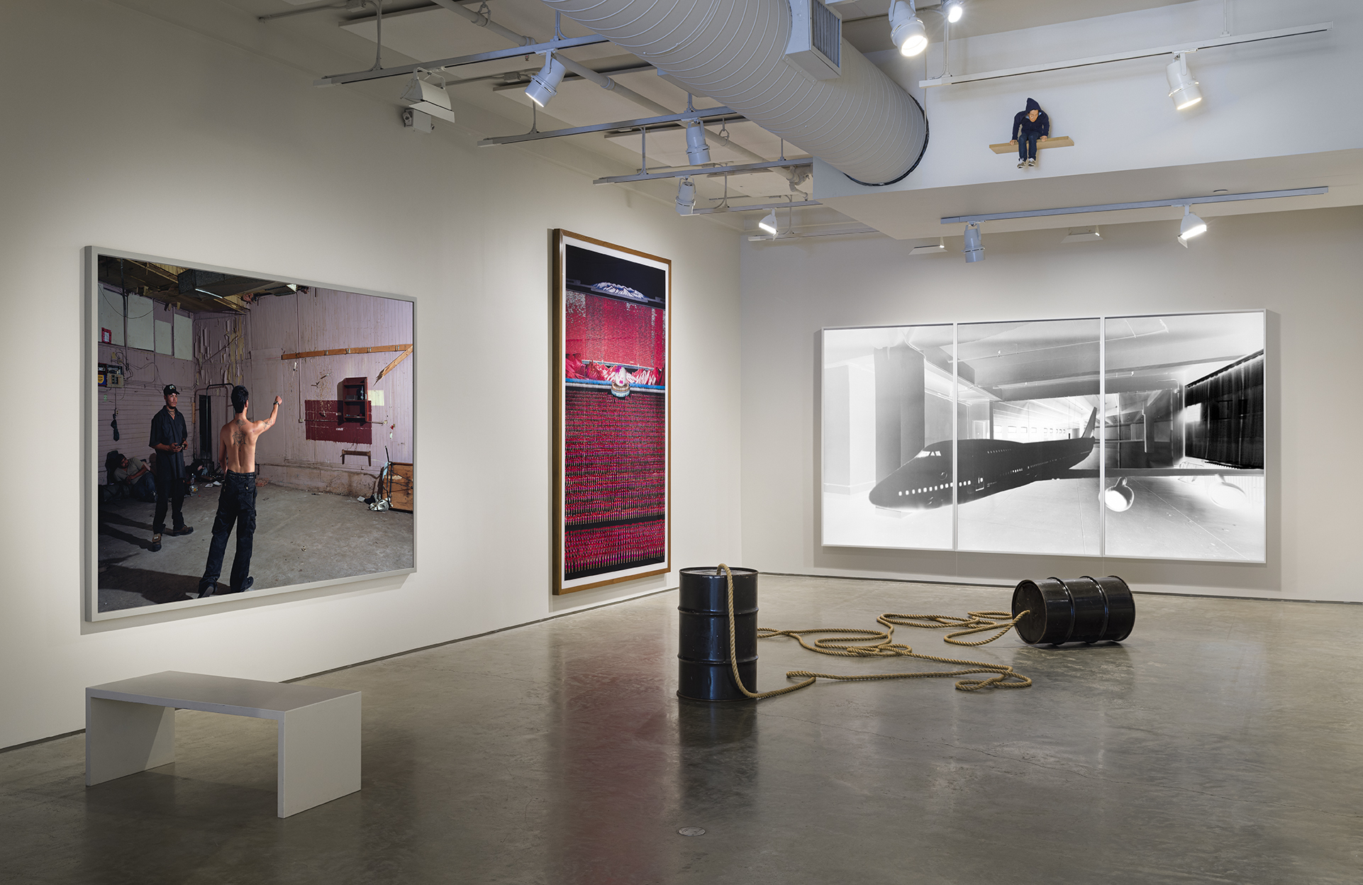 From left to right: Jeff Wall, Knife Throw, 2008, reprinted 2019. Color photograph, 75 3/8 x 104 x 2 inches; Andreas Gursky, Pyongyang IV, 2007. C-print, 82 5/16 x 101 15/16 inches; Vera Lutter, Studio, XXX: June 29 - July 18, 2006, 2006. Unique silver gelatin print, 91 1/2 x 168 inches. Center: Christian Marclay, Dialogue of the Giants, 1998. Metal drums and rope, each Drum: 28 3/4 x 18 1/2 inches. Photo by Etienne Frossard.