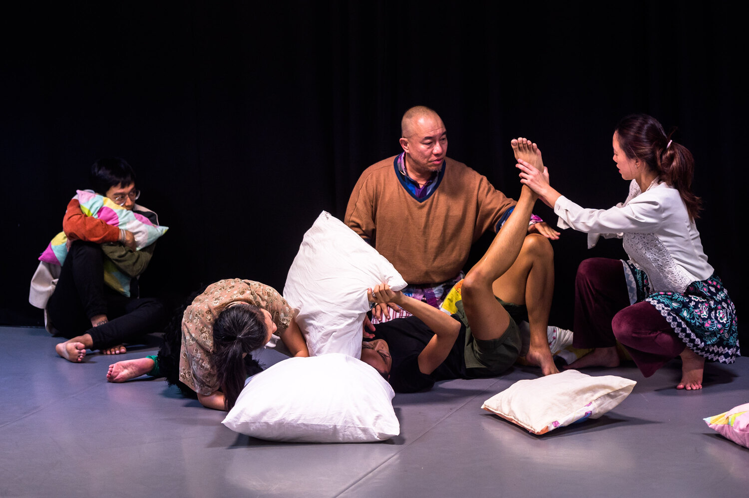 IMAGE DESCRIPTION: There are pillows and people and plenty of colours and texture. Looking more closely, on one side there are 4 people touching each other’s limbs and bodies gently, seemingly forming one larger connected body through having their hands on someone else’s ankle or knee. On the other side, a person hugs a pillow tightly while watching the other 4.