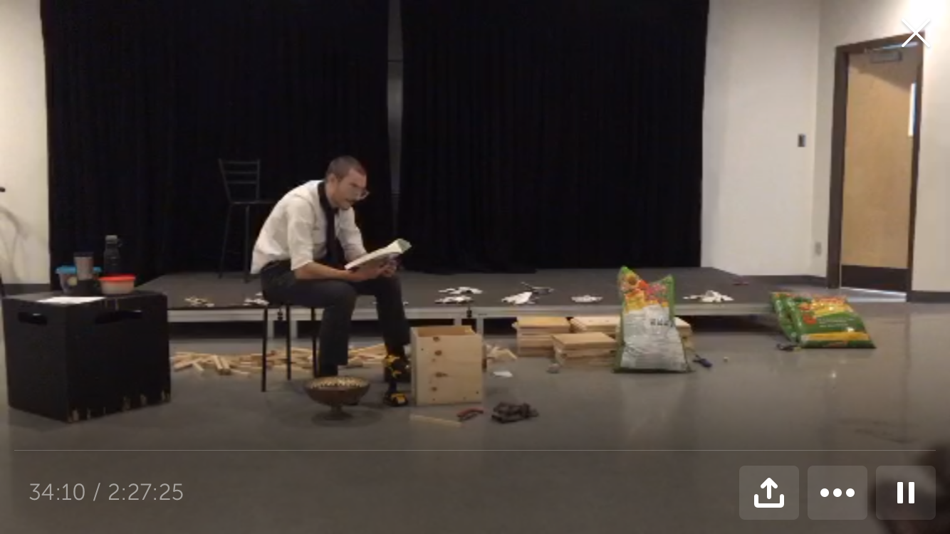 IMG Description: Screenshot from a livestreamed video. Fargo, dressed in a white button down shirt and dark slacks, sits on a stool reading from a book. A wooden box is at his feet. Behind him is a small stage with shards of plates on it; he is surrounded by pieces of wood and two bags of soil. Next to him is a black wooden cube with a water bottle and containers of food on it.