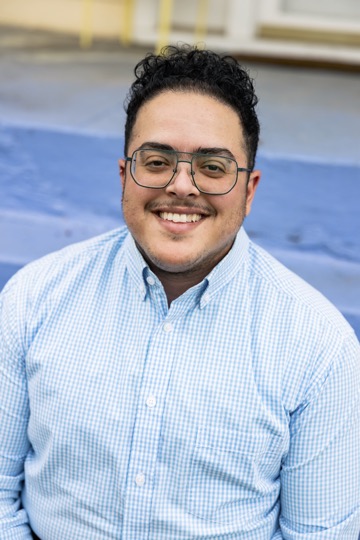 Image description: Aimi Hamraie smiles at the camera, wearing a cheerful blue checkered shirt.