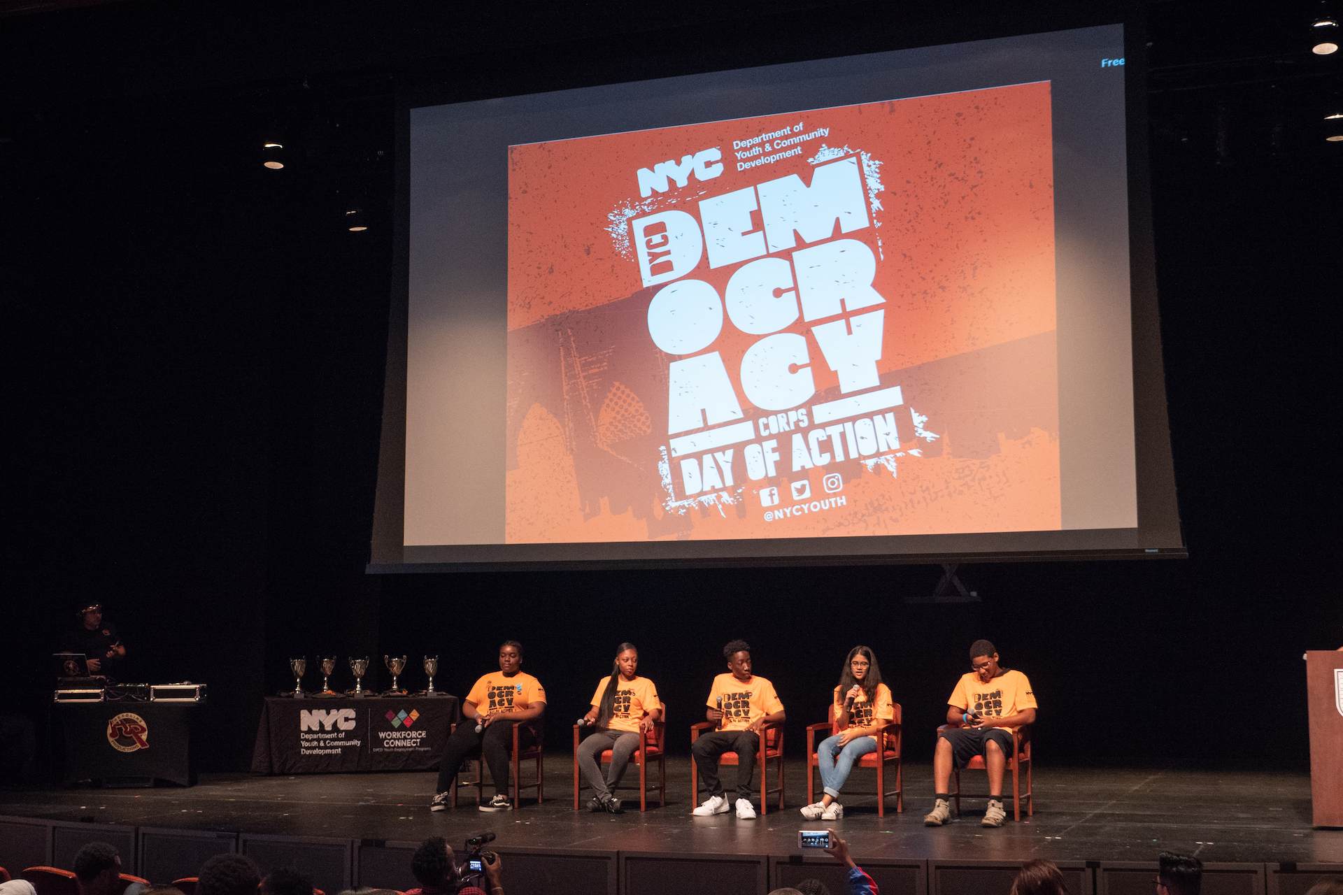 A panel for five Summer Youth Employment Program participants on a stage wearing neon orange t-shirts with the Democracy Corps logo in one color black. Behind the youth participants is a large screen protection of the Democracy Corps logo in white on top of a orange distressed New York City cityscape background.