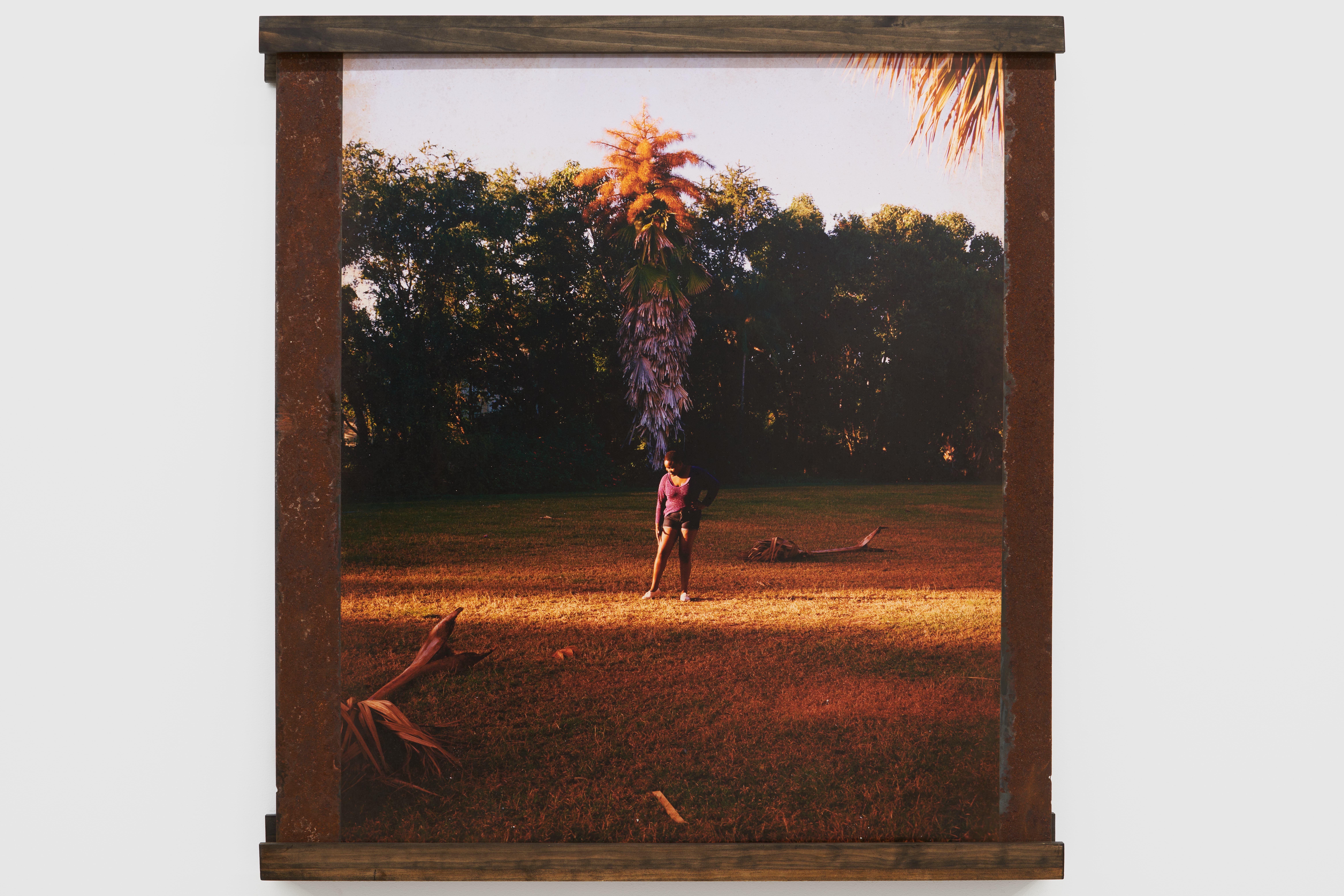 A partially framed color photograph printed on a rusty metal substrate hangs on a white wall and depicts a lone person from far away standing in a clearing, surrounded by a line of tall trees at dusk.