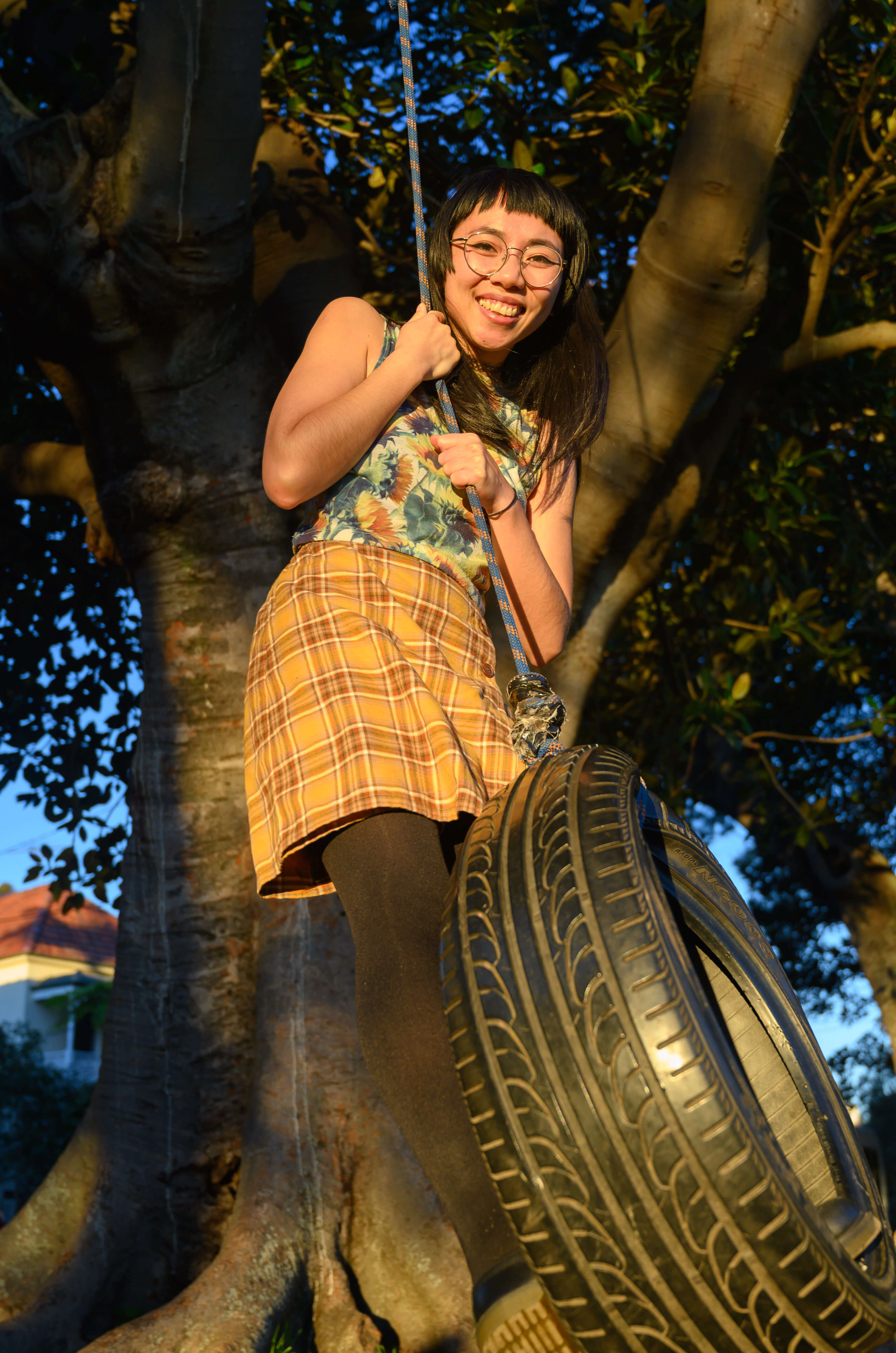 Annie holds onto a tire swing and gazes down on the camera. They have a vibrant smile on their face.