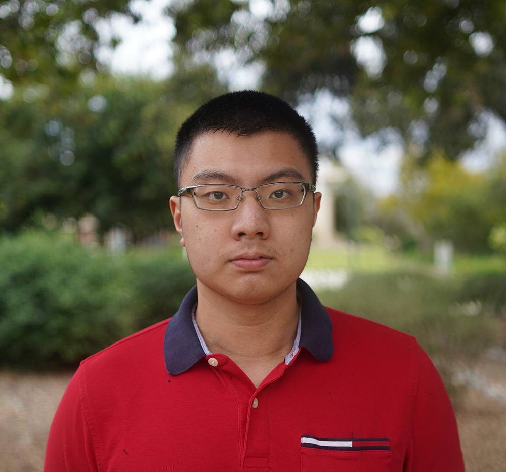 A headshot of a person in a crew cut, glasses, and a polo shirt against a blurred background of trees.