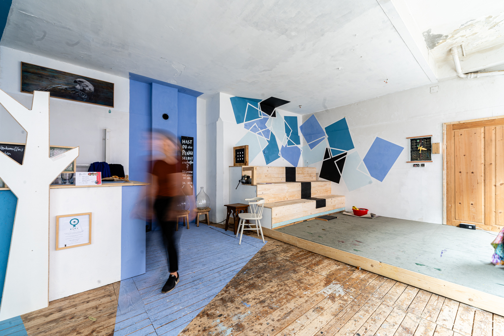 Bild 5 Cafe Eder: Das Bild zeigt wie Bild 4 einen Ausschnitt der Sitzecke des B&uuml;hnenbereichs, sowie das rechte Ende der Bar. Ein Teil der Wand ist zwischen Bar und Sitzbereich Lila, die Farbe verl&auml;uft am Boden  zu einem Spitz zusammen. Der restliche Boden ist aus Holzdielen.