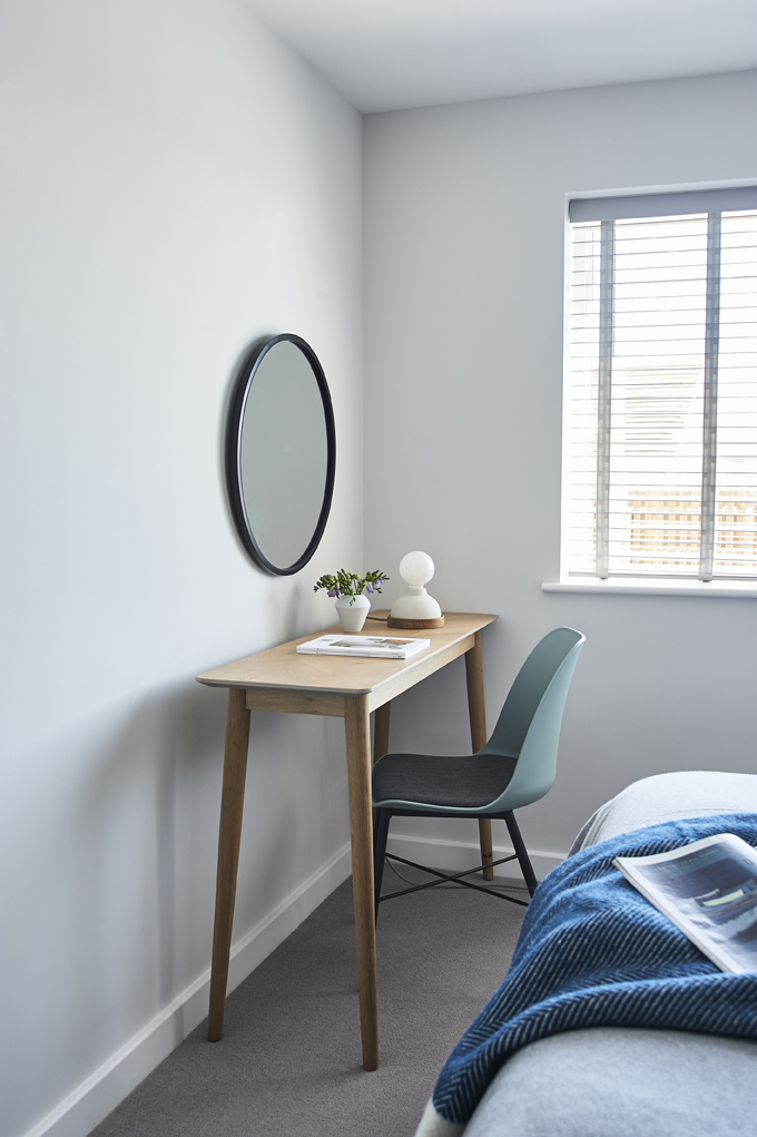 Close-up of wooden desk with teal chair, pot plant, magazine and small lamp. Round mirror on white wall.