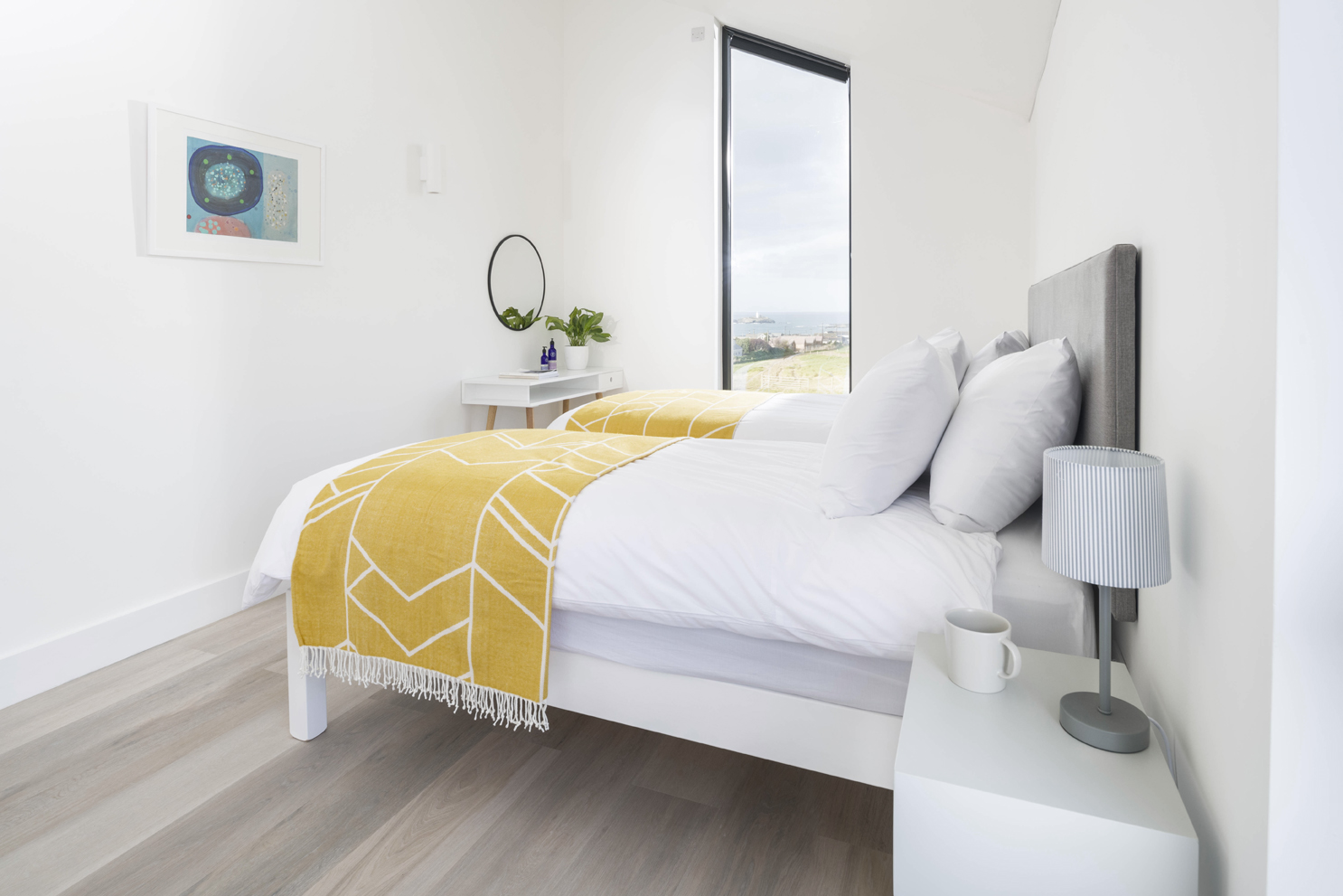Bright, modern bedroom with double bed. Grey upholstered headrest with cushions and yellow throw. White desk and side table. 