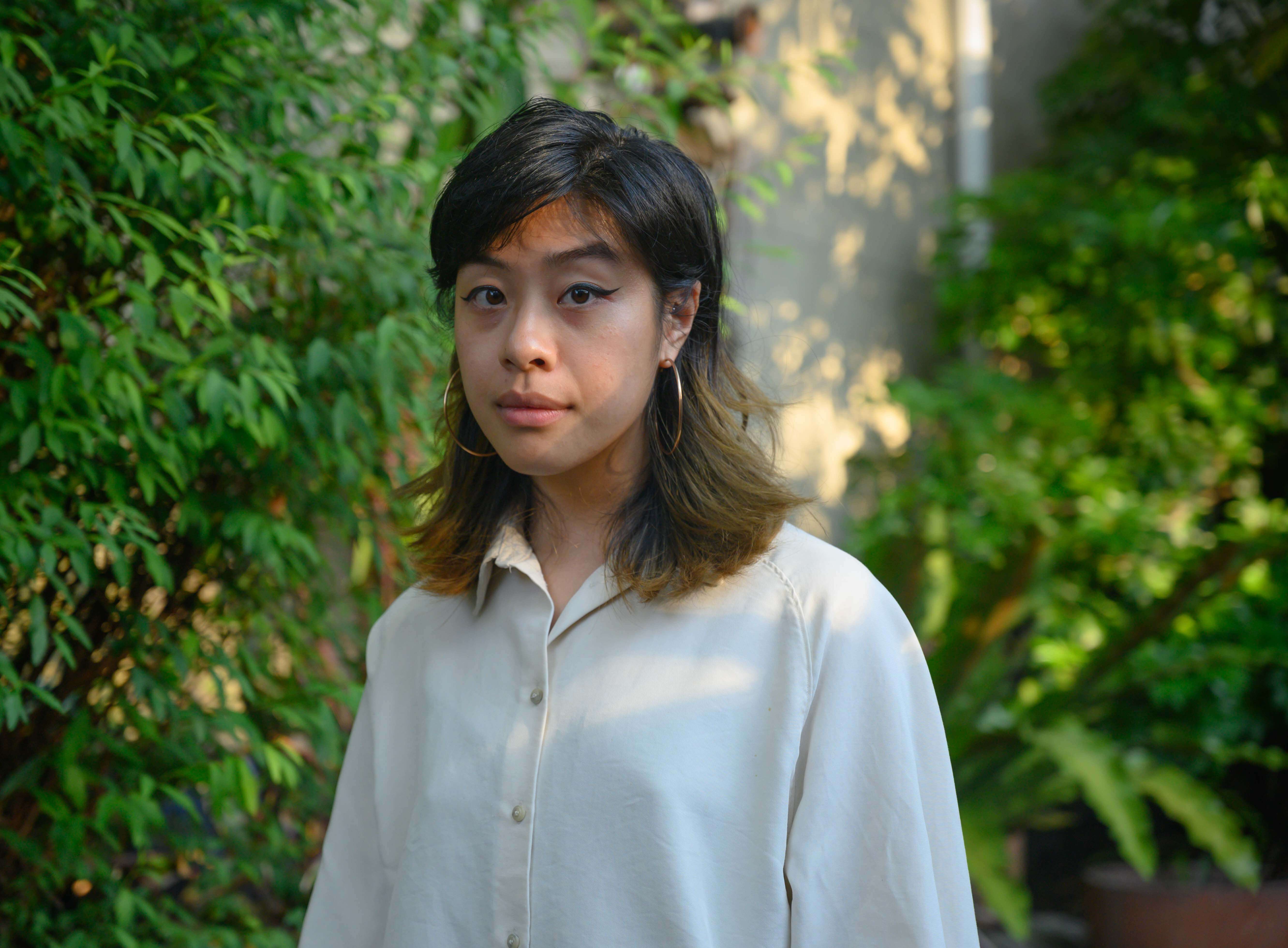 Grace, a light skinned Asian woman, stands in front of a leafy wall. Pictured from her chest up and she looks directly at the camera.