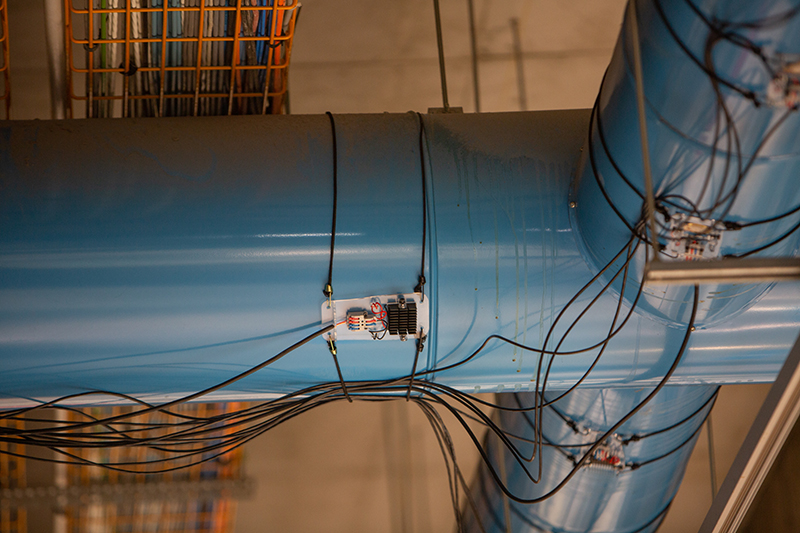 Blue pipe with wires strapped to it and attached to electronic devices. Concrete and a red metal grate in the background.