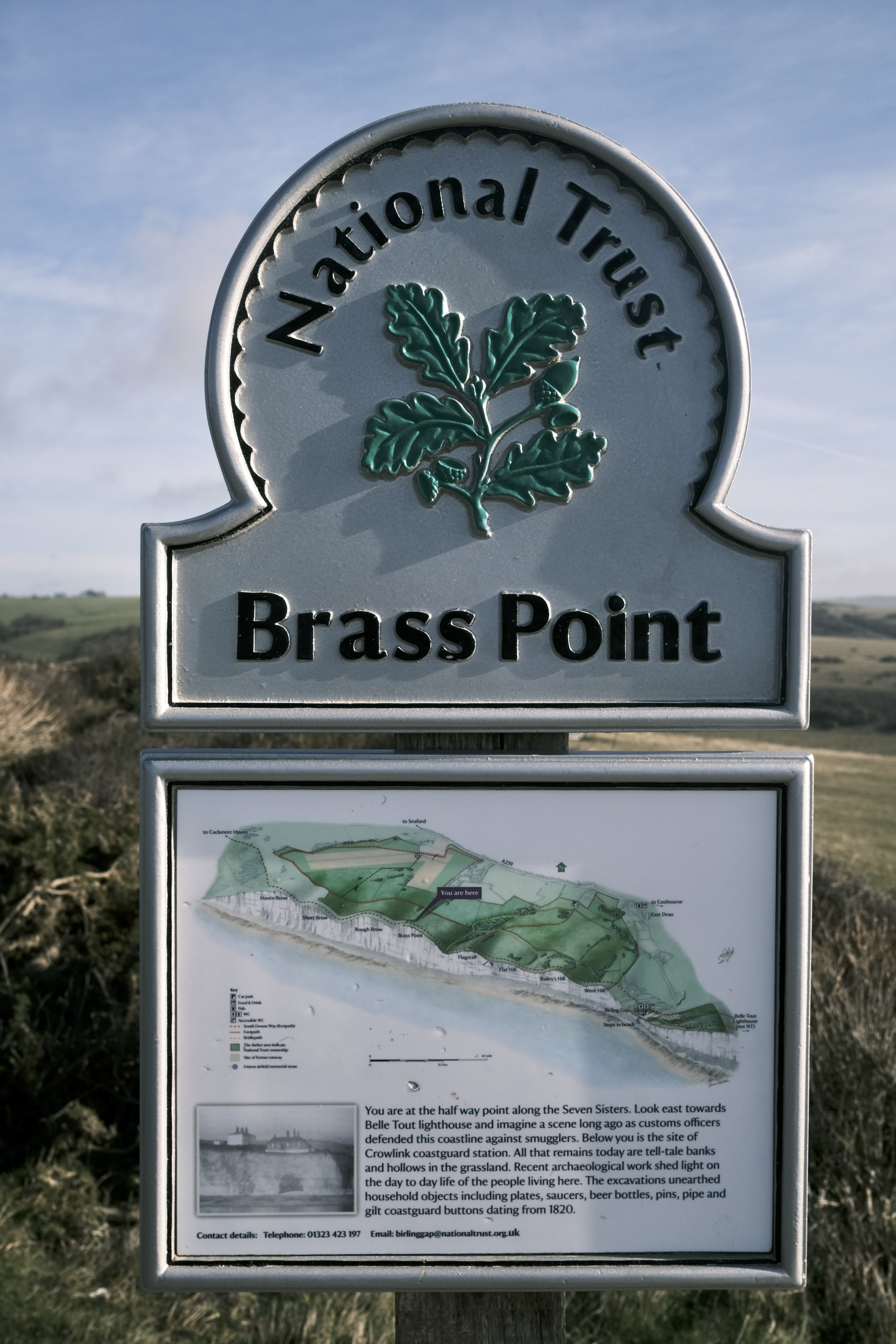 A National Trust marker for Brass point. It shows a map of the river area and tells people they are at the halfway point of Seven Sisters. Green hills and a blue sky with some light white clouds can be seen beyond the marker.