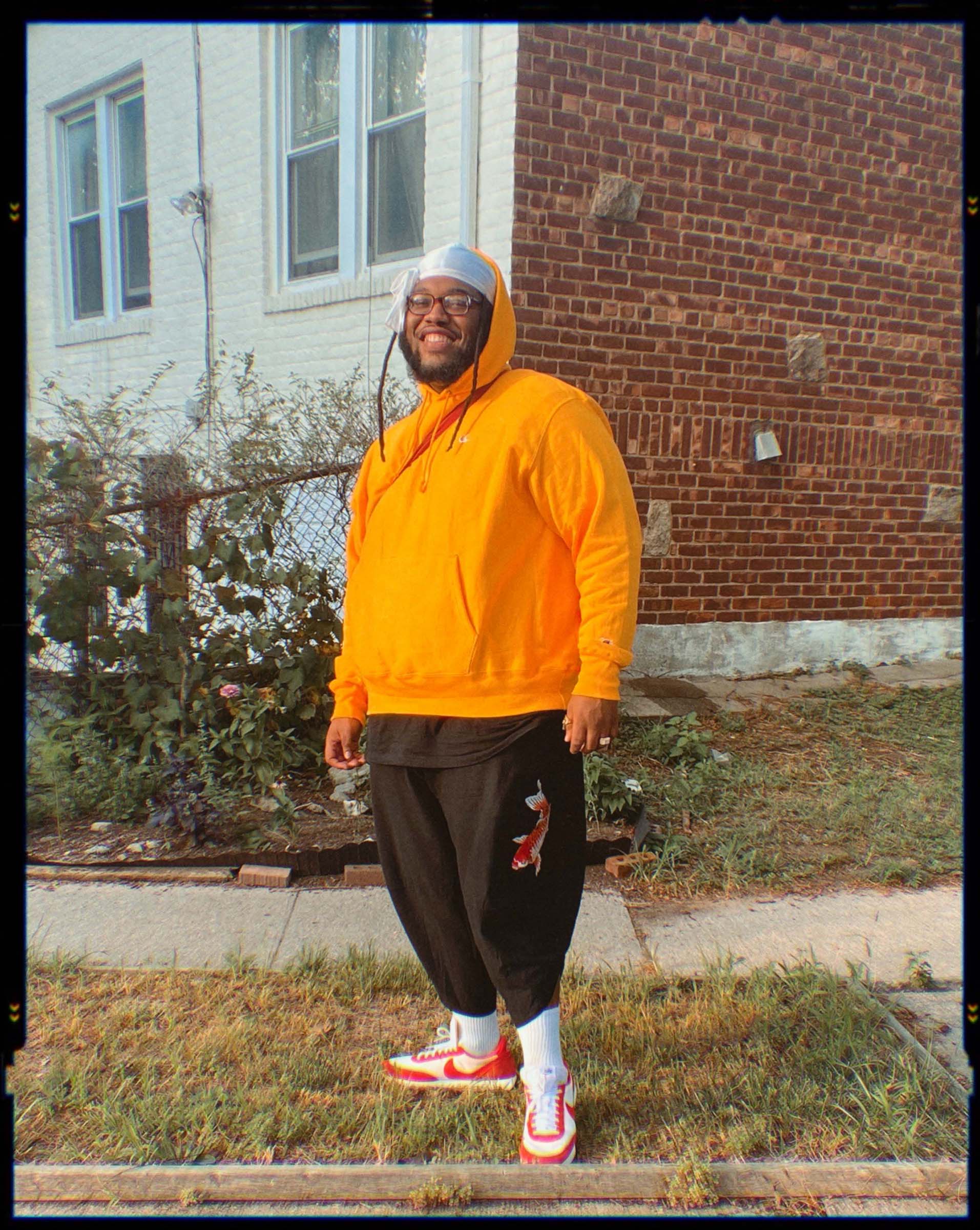 Jeffrey Dean Chambers wearing a bright orange hoodie, white cap and trainers stands on the verge of a residential street.