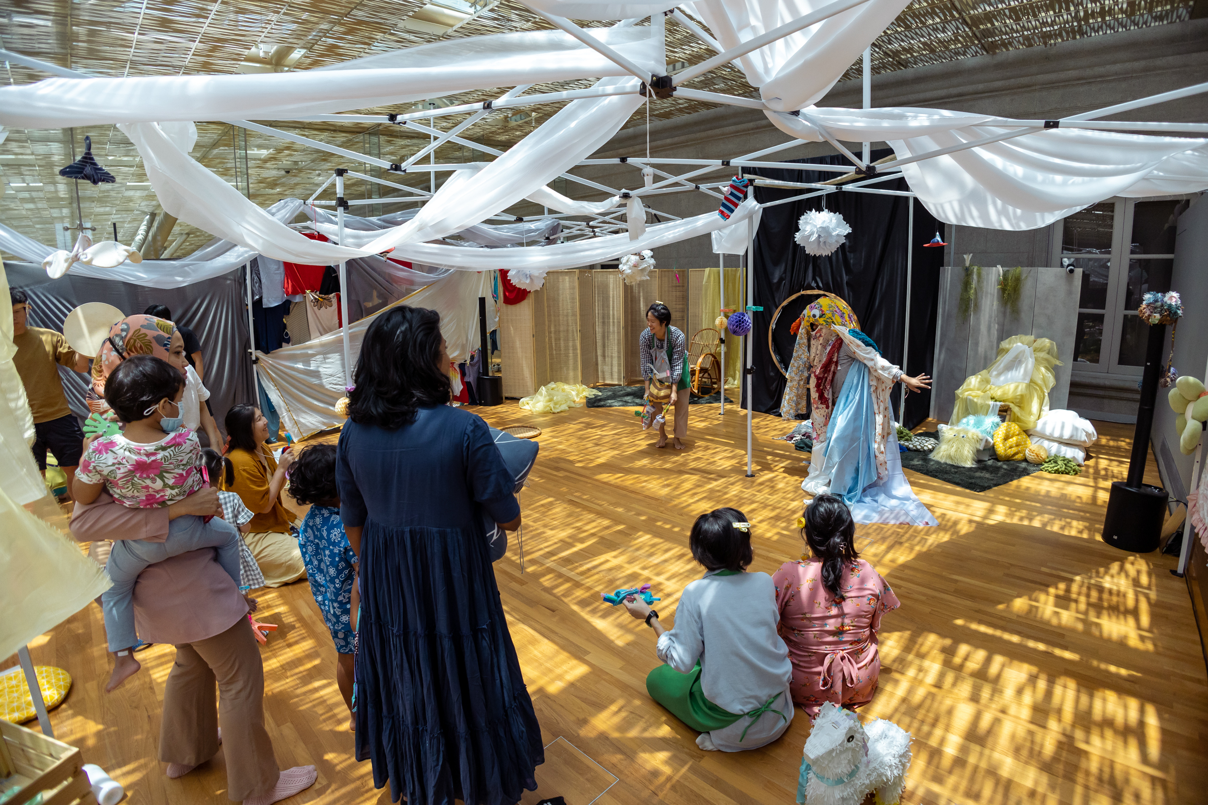 IMAGE DESCRIPTION: A wide shot of the performance space, which is set up in a whimsical fashion - there is a canopy of narrow white fabrics draped around structures like a soft sky and things hanging down that look like clouds or birds, while the floor is covered in a weave-like pattern because of the shadow cast from the skylight. Young children and their parents are scattered around the space, some standing and some sitting, looking at two performers, one of whom is dressed as a creature with an extra large head, large googly eyes, and whose body is made of overlapping colourful fabrics.
