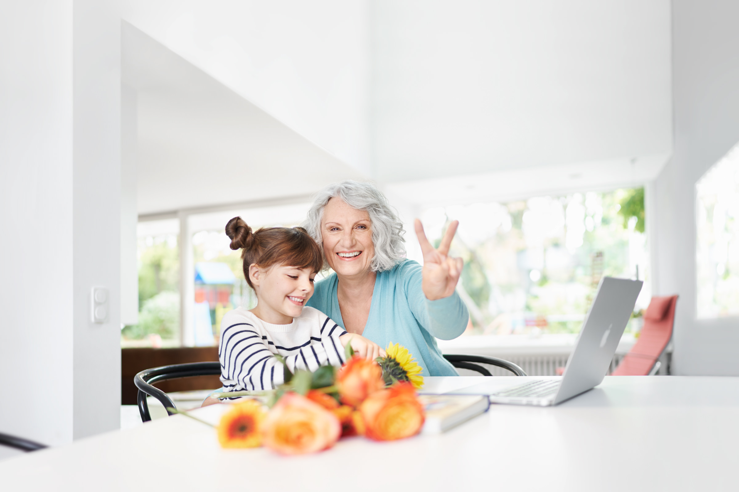 Portrait einer Oma mit ihrer Enkelin, sitzend am Esstisch vor einem Laptop. Die Oma zeigt das Victory-Zeichen in die Kamera.