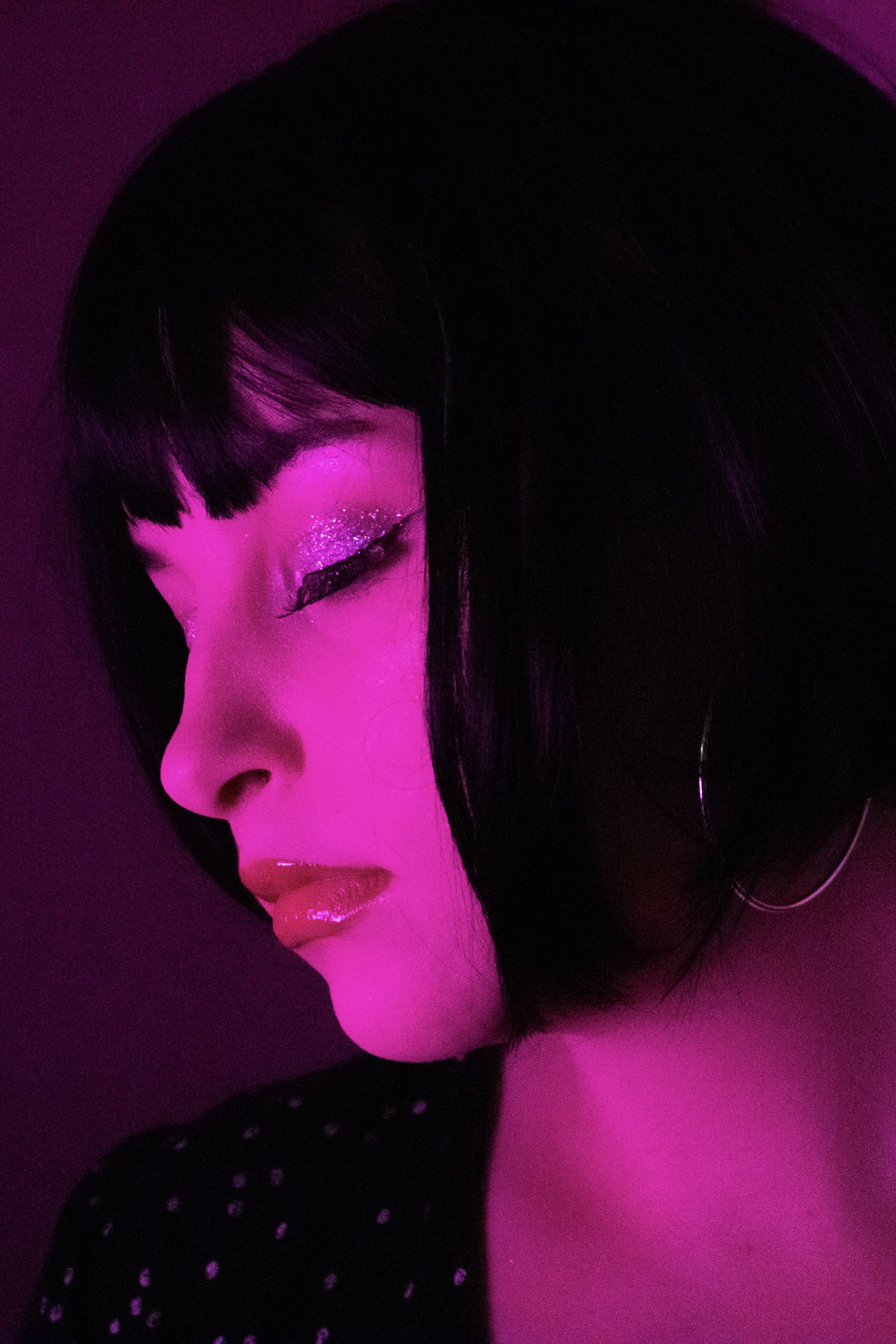 Close-up portrait of a woman with a sharp black bob and shimmering sparkly eyelids, illuminated in high-contrast neon pink light. A mysterious, eerie yet fashion-forward aesthetic.